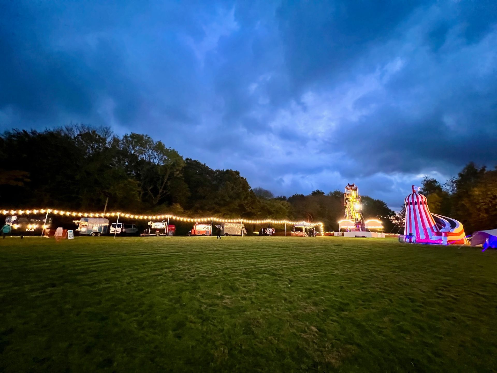 Carnival at dusk: food stalls, rides, and a slide on a grassy field beneath a cloudy sky.