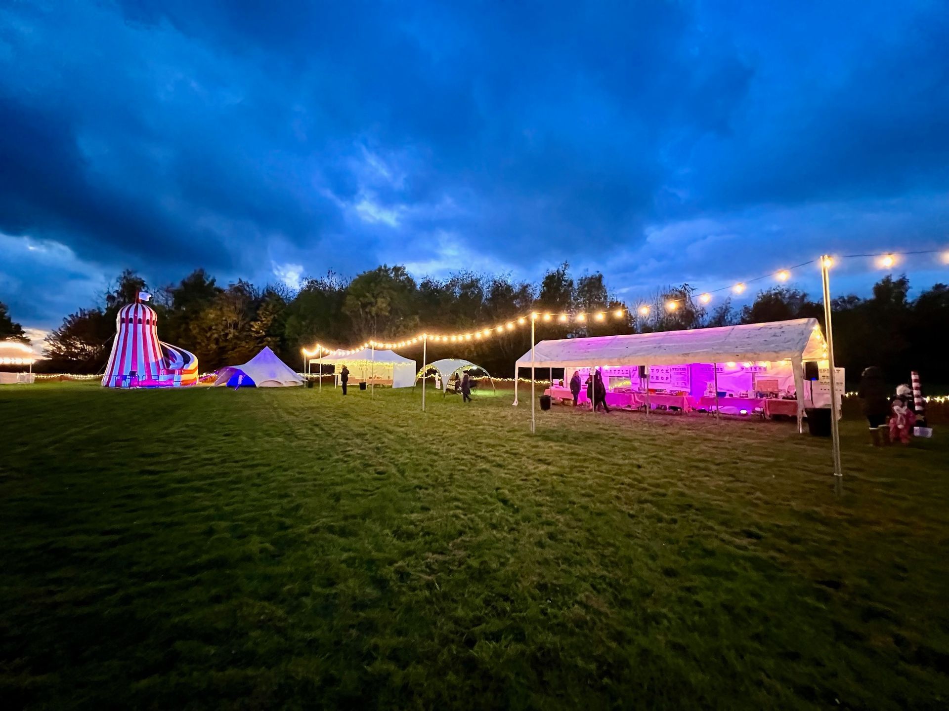 A nighttime festival scene with tents, string lights, and a colorful carnival ride on a grassy field.