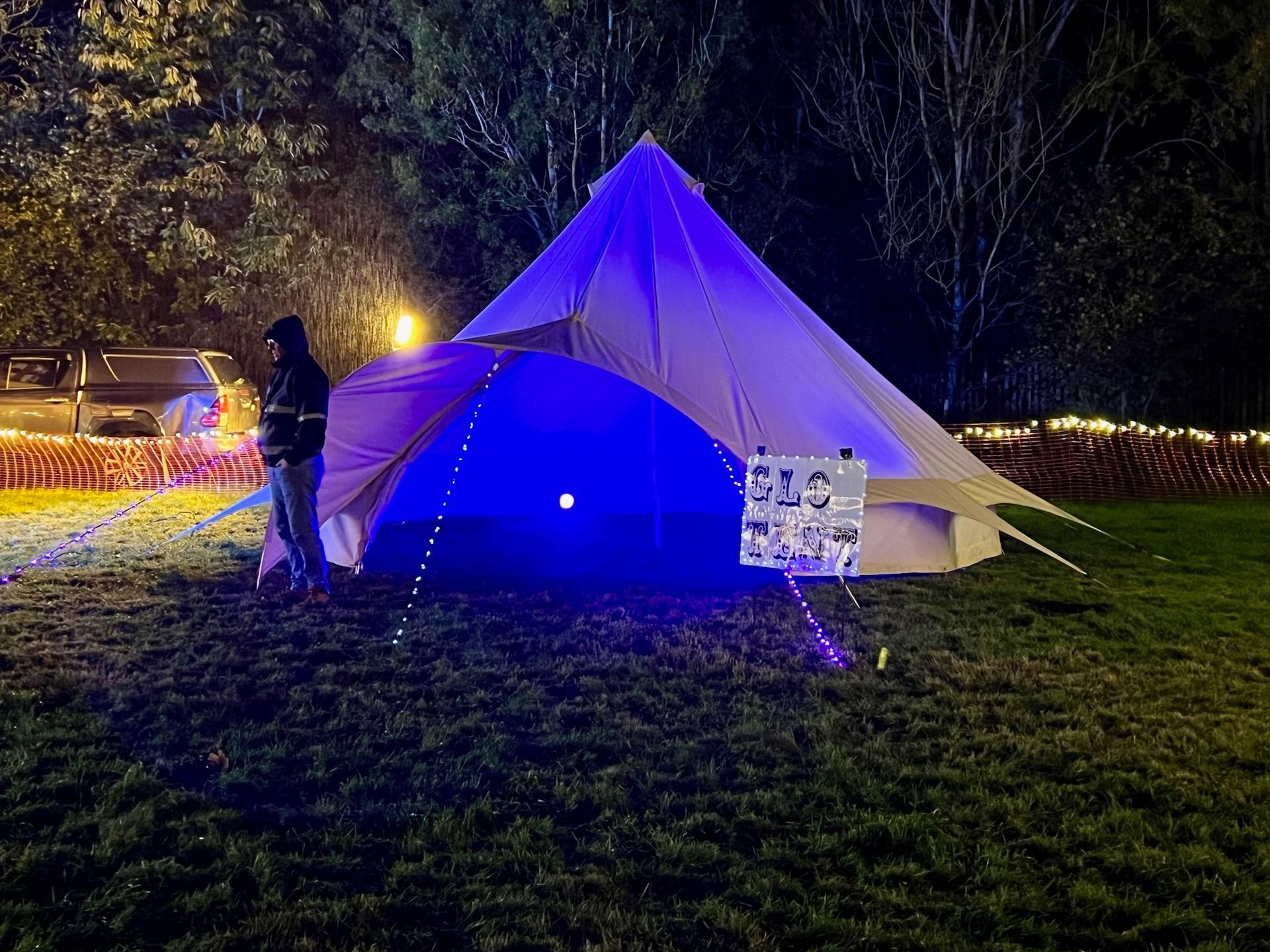 Person stands near a lit tent at night; blue lights, sign, and a vehicle in the background.
