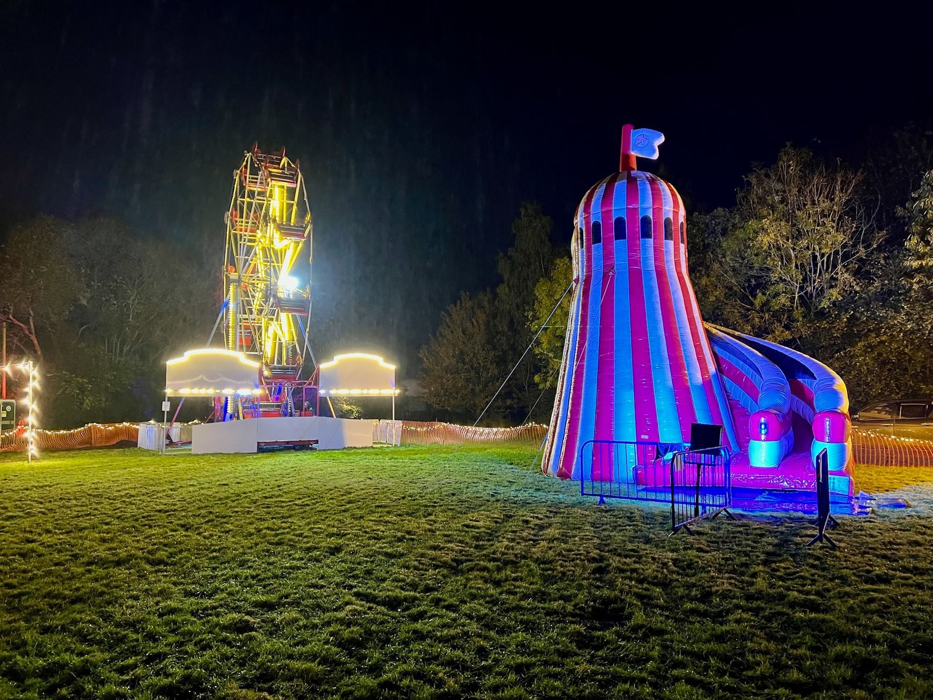 A nighttime carnival scene with a lit Ferris wheel, colorful inflatable slide, and grassy lawn.