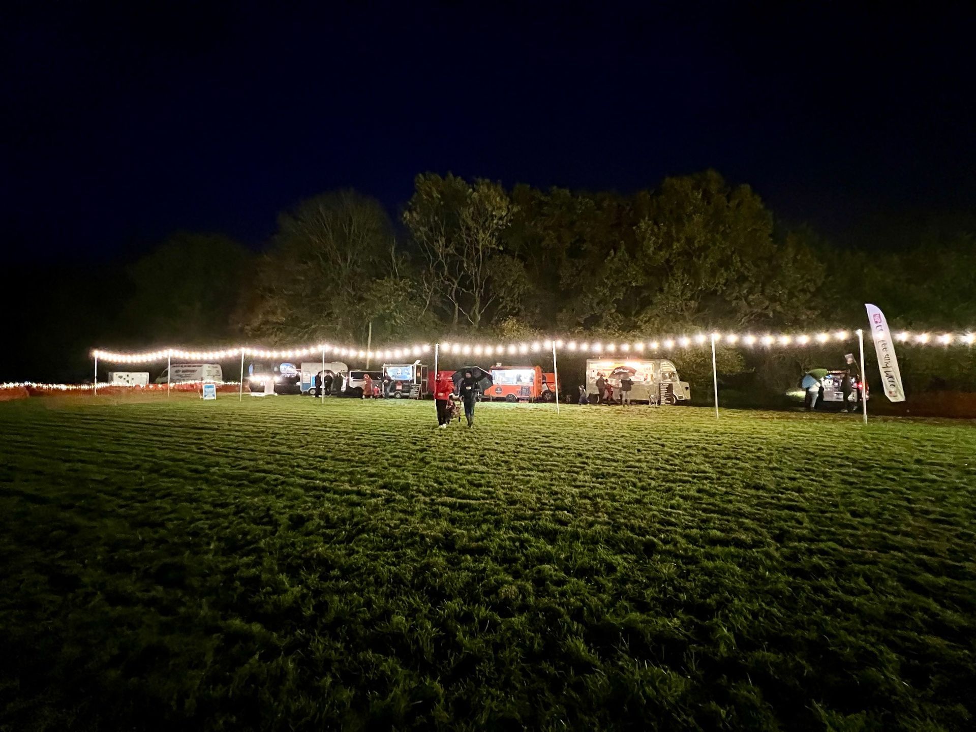 A nighttime outdoor event with string lights above food trucks and a grassy field; people are present.
