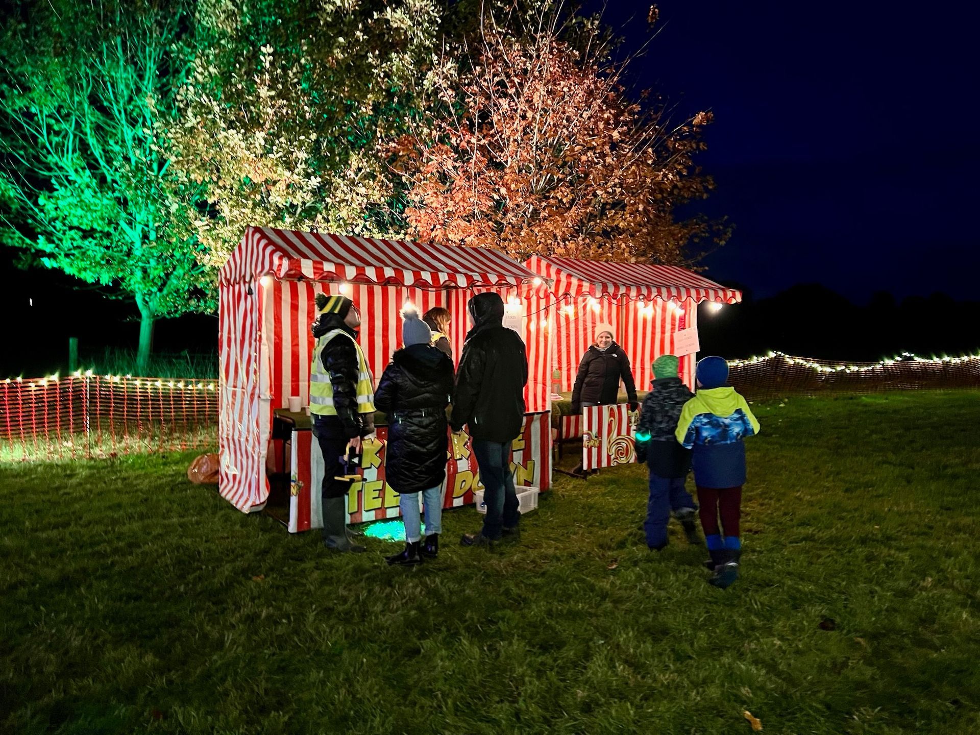 People at a Christmas market stall lit by red and green lights at night.