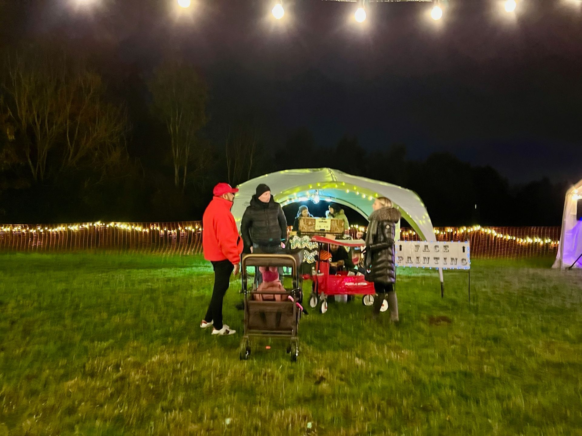People stand around a lit-up food cart in a grassy field at night; string lights and a small fence are visible.