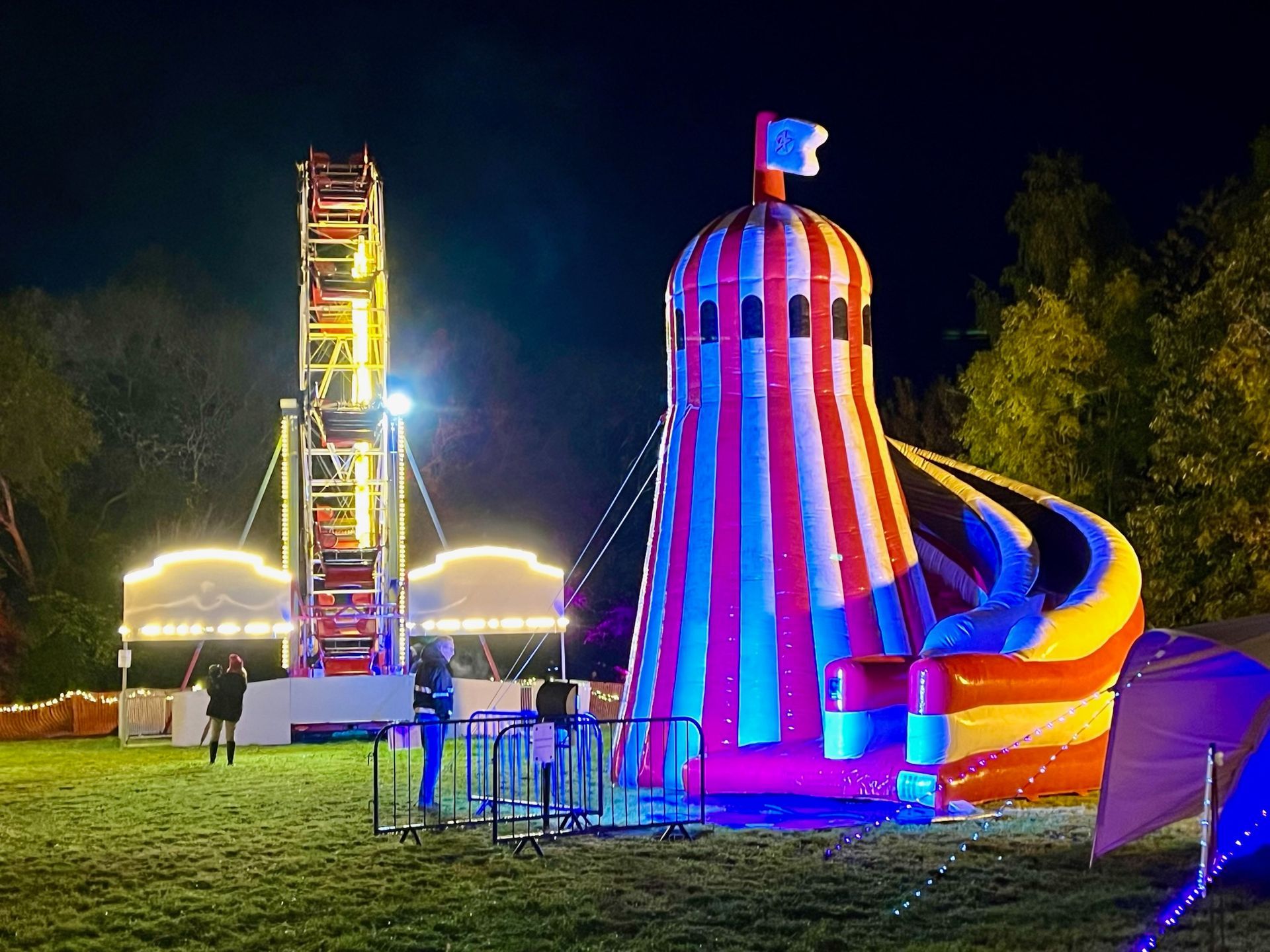 A brightly lit carnival at night with a Ferris wheel and inflatable slide on a grassy field.