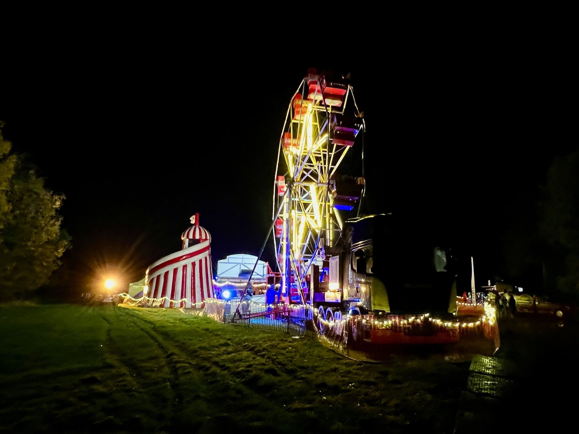 Ferris wheel and carnival games illuminated at night on a grassy field.