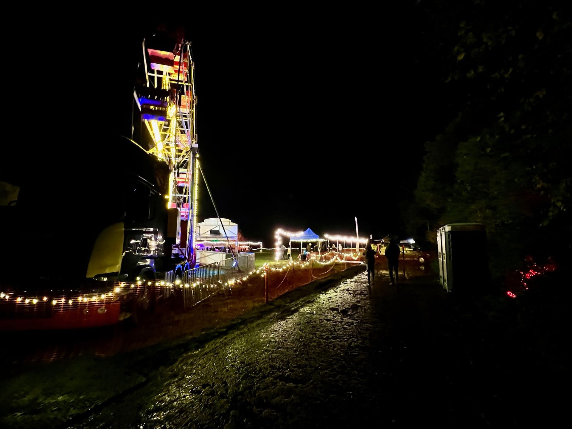 Nighttime view of a lit carnival with a Ferris wheel. People walk along a pathway.