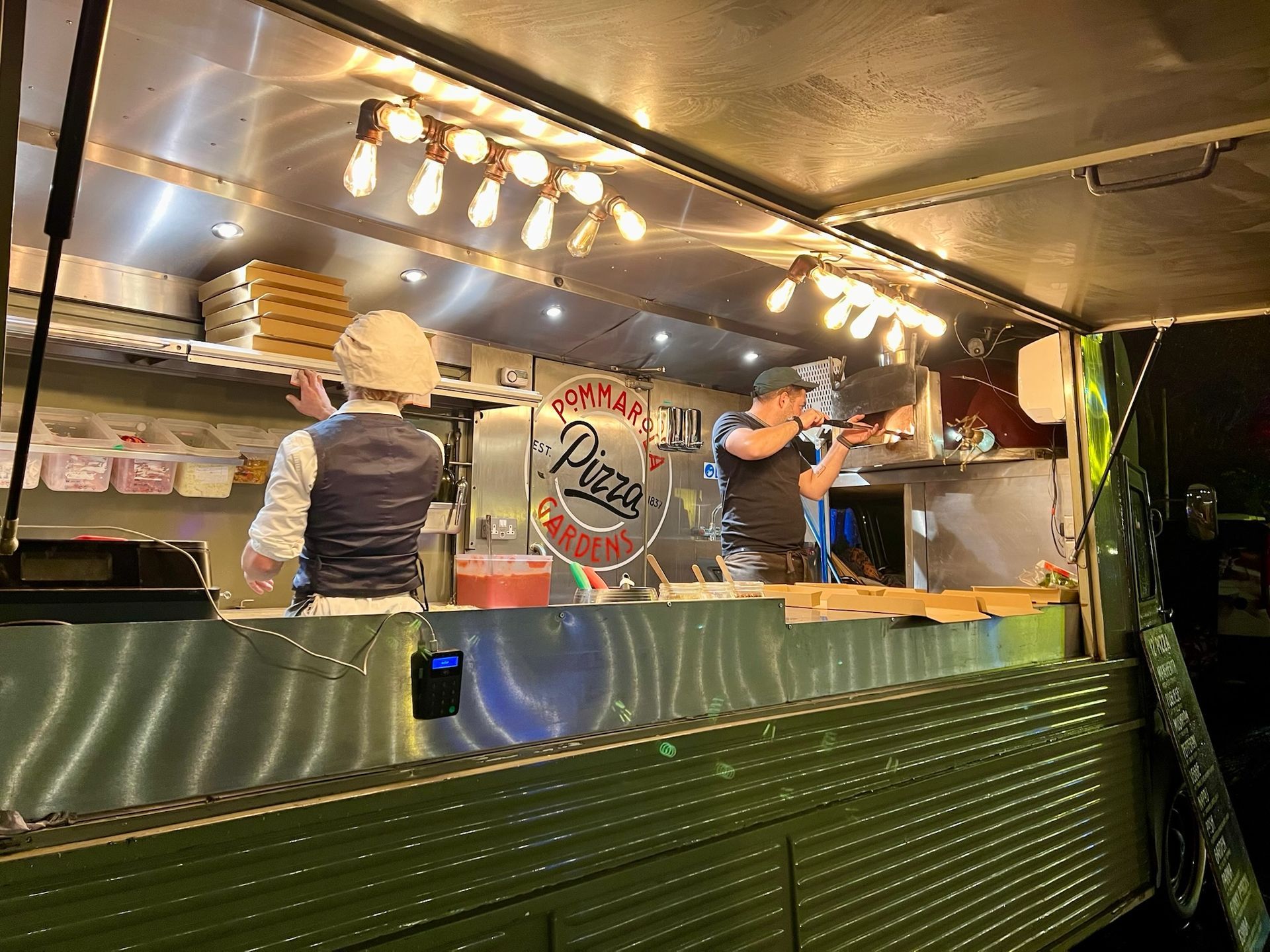 Two people working inside a pizza food truck. One prepares dough, the other takes an order. Green exterior, string lights.