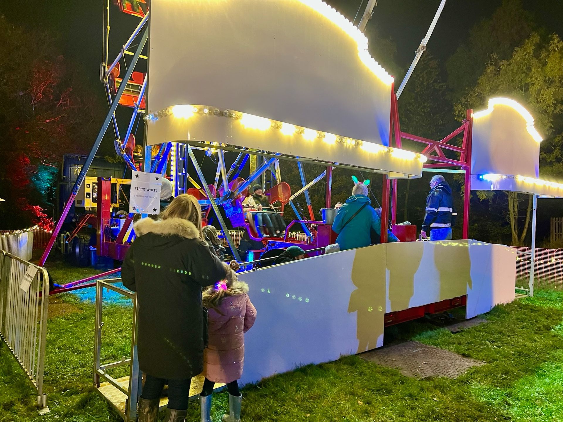 A Ferris wheel ride at night. A mother and child in coats are standing near the ride.