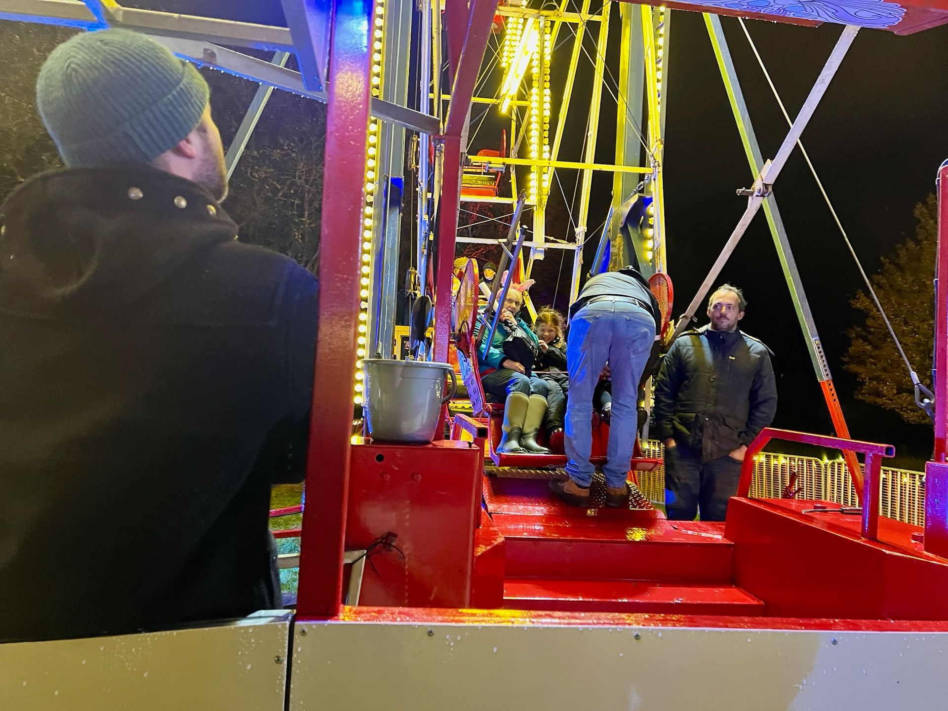 People on a carnival ride at night, red and white structure, lights, one person in a green hat.