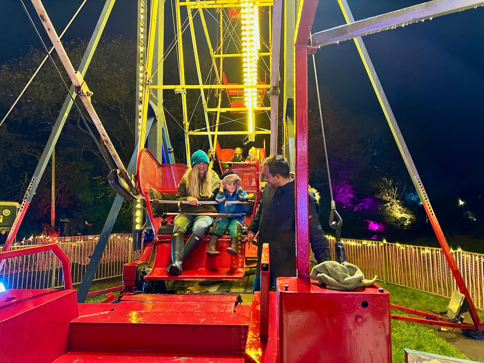 Family on a brightly lit amusement park ride at night. Child smiles between seated adults. Red platform, yellow lights.