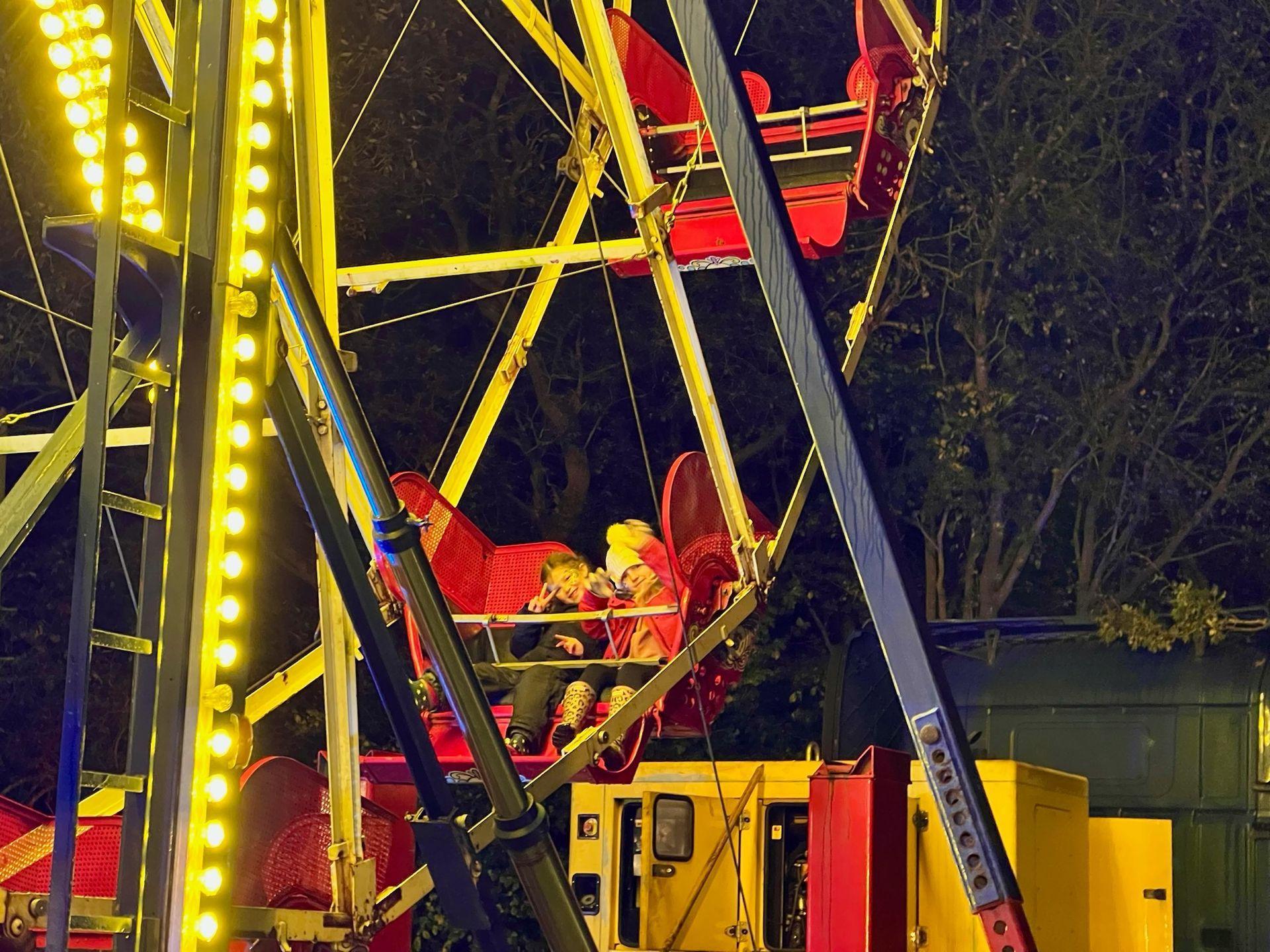 Ferris wheel at night, illuminated with yellow lights, carrying passengers in red cars.