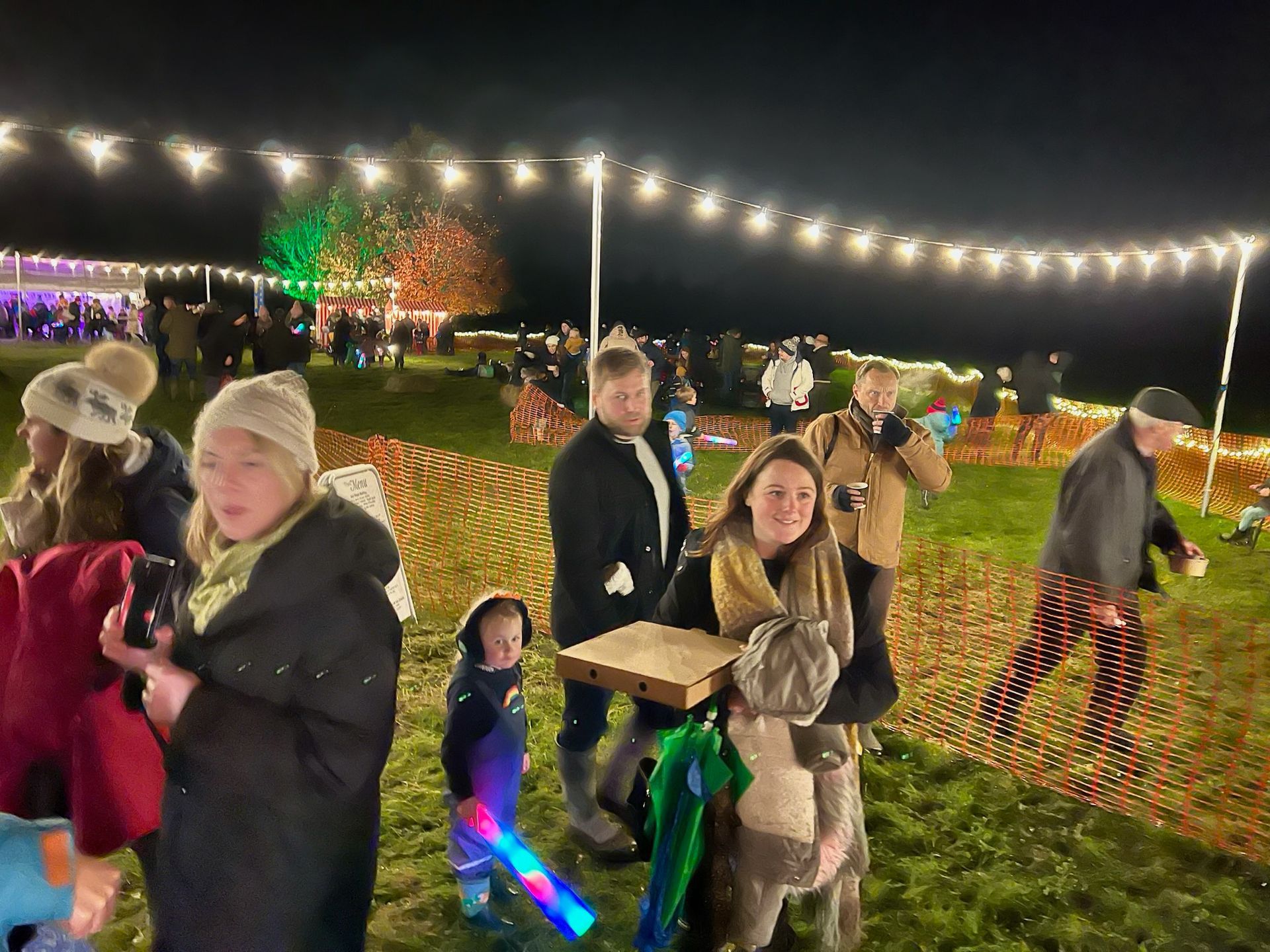 People at a night event, some holding phones, food, or glowing toys. String lights and illuminated trees in the background.