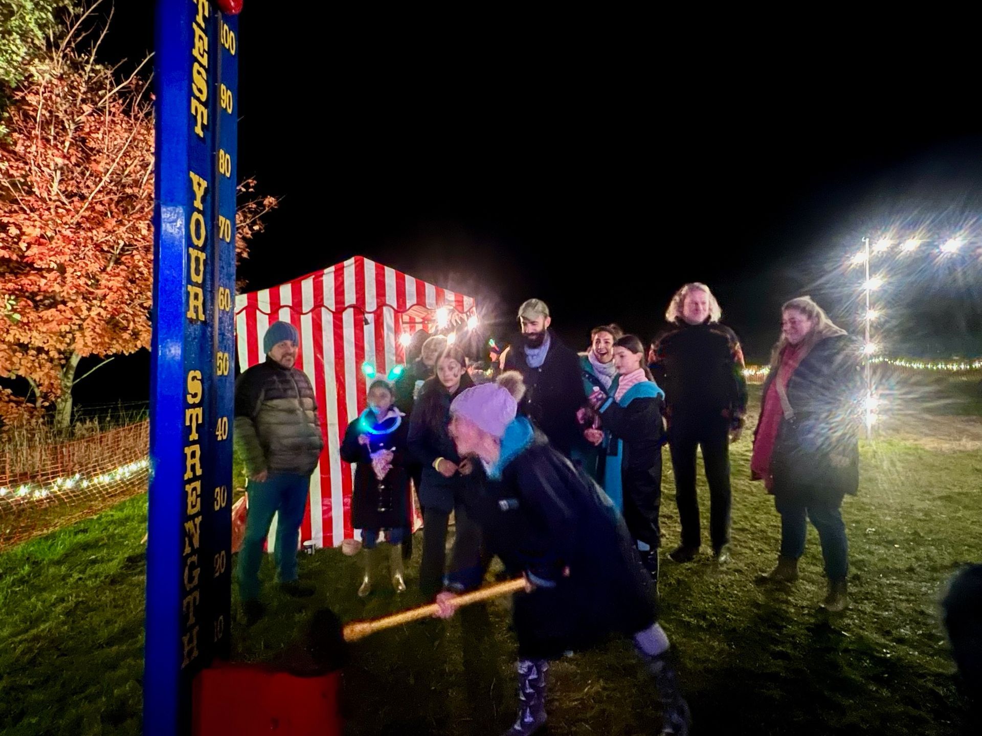 Person swings a hammer at a strength test game at night. Others watch, in front of a striped booth.