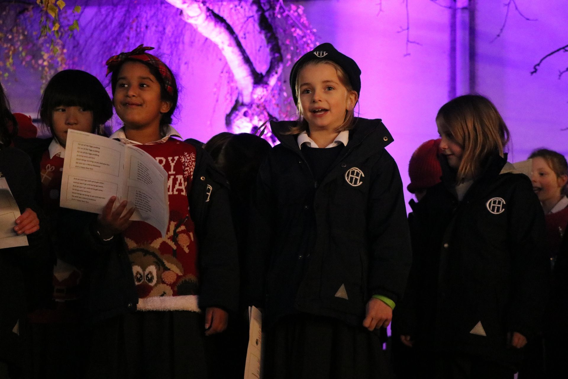 Children singing carols outdoors under purple stage lighting.