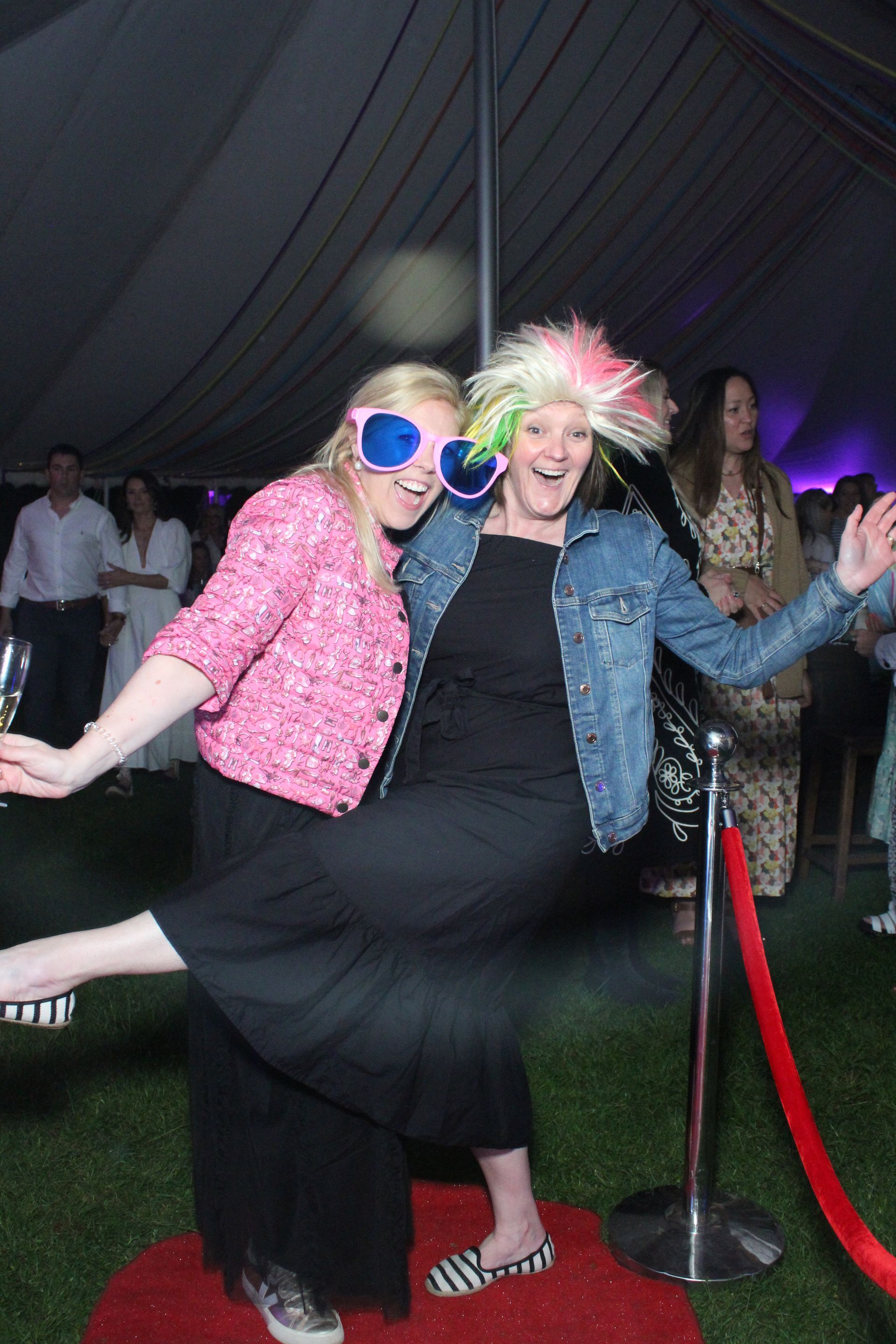 Two women pose on a red carpet. One in a pink jacket and large glasses, the other in a wig and denim jacket.