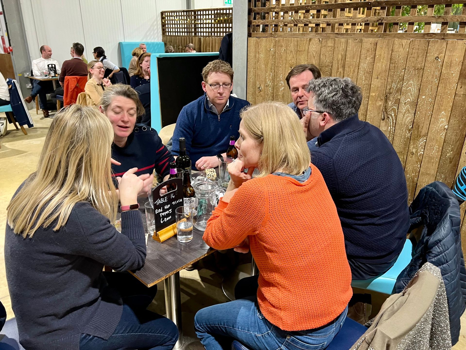 Group of people in a casual setting at a table, conversing.  Orange, blue, and grey clothing.  Indoors.