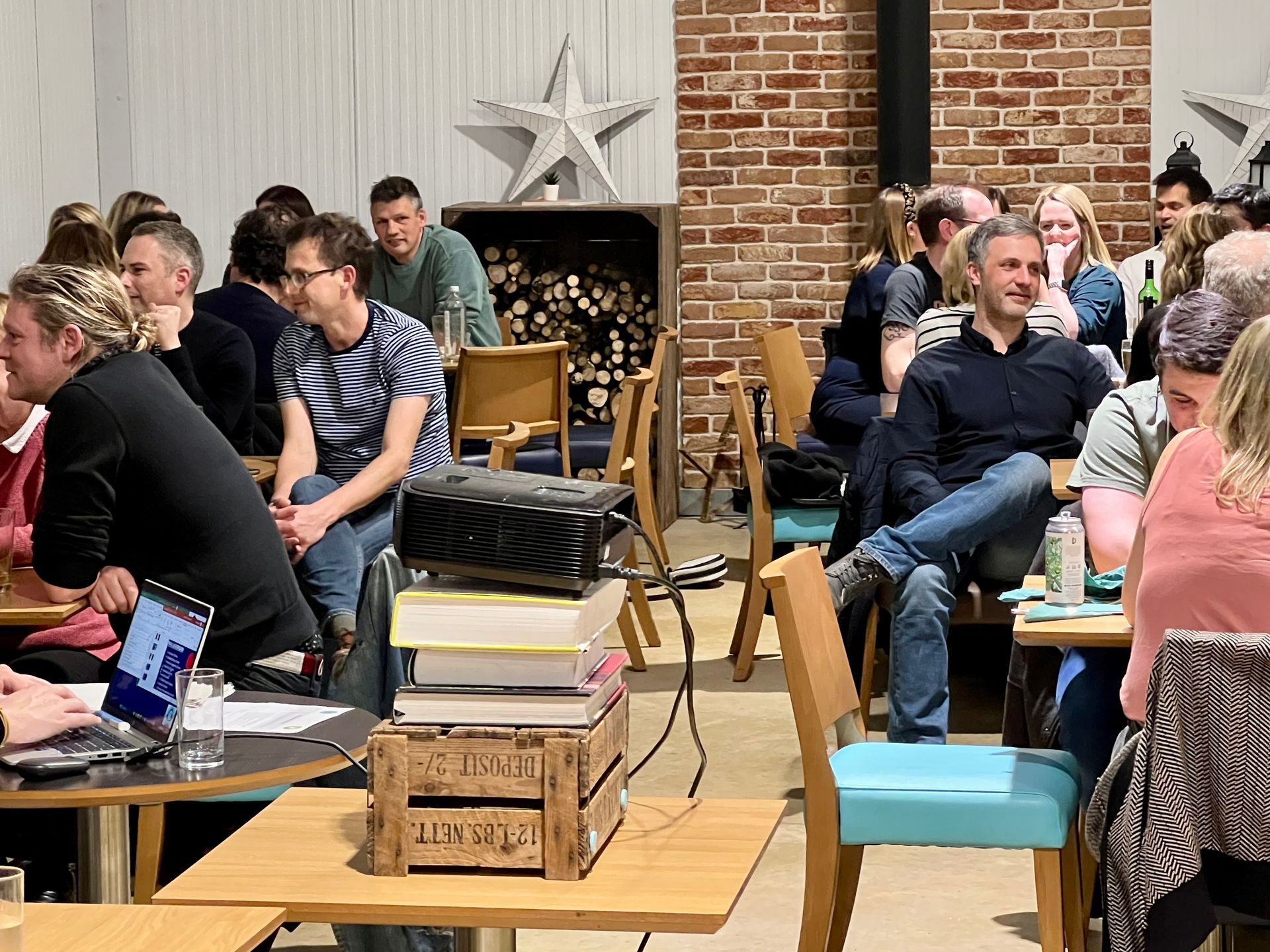 People in a cafe listening to a presentation. A projector sits on stacked books. Brick wall and fireplace in background.