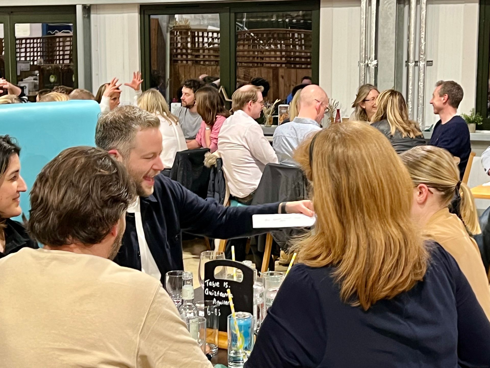 People at tables in a restaurant, chatting and smiling. Some are raising their hands, suggesting a lively atmosphere.