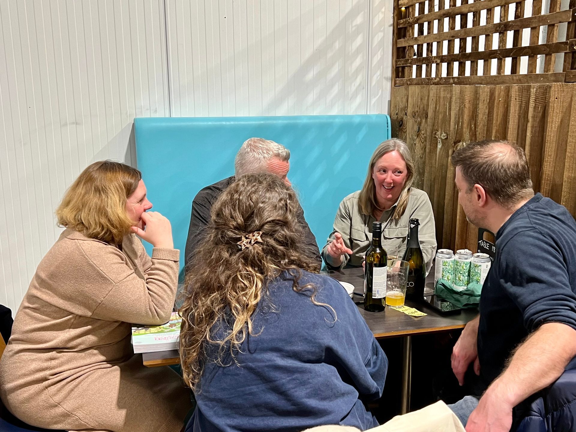 Five people seated at a table, conversing. One woman gestures, bottles, cans, and snacks visible. Rustic setting.