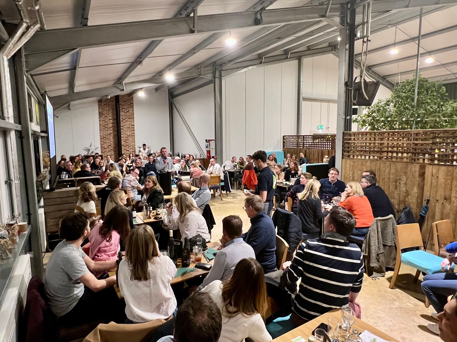 Large indoor gathering with people at tables, some standing and talking, with a speaker in the center.