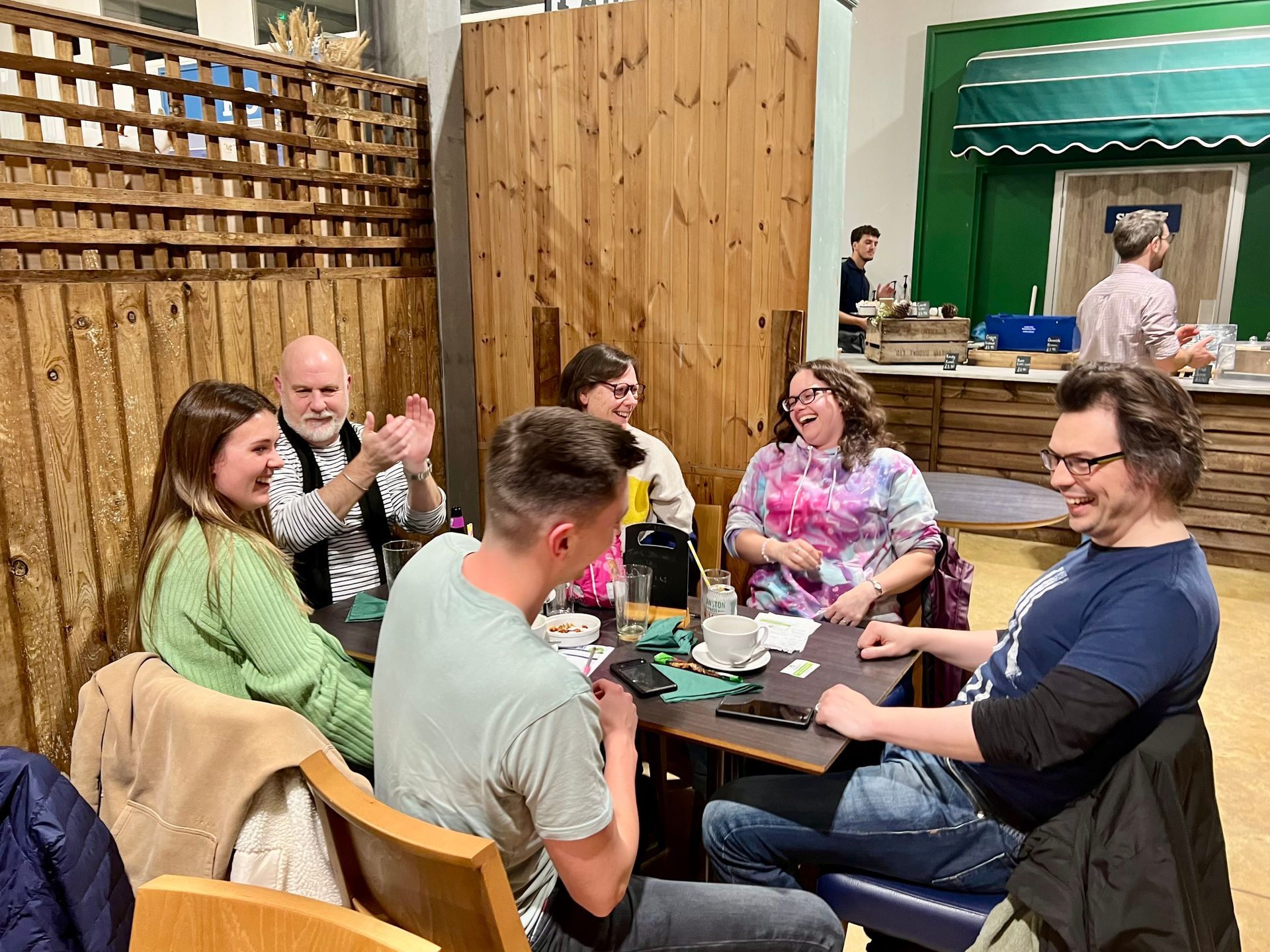 Group of people laughing around a table in a warmly lit room with wood paneling.