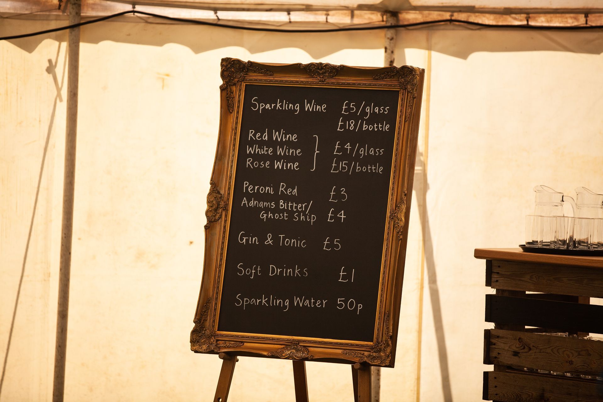 A chalkboard menu on an easel displays drink prices; a tan tent is in the background.