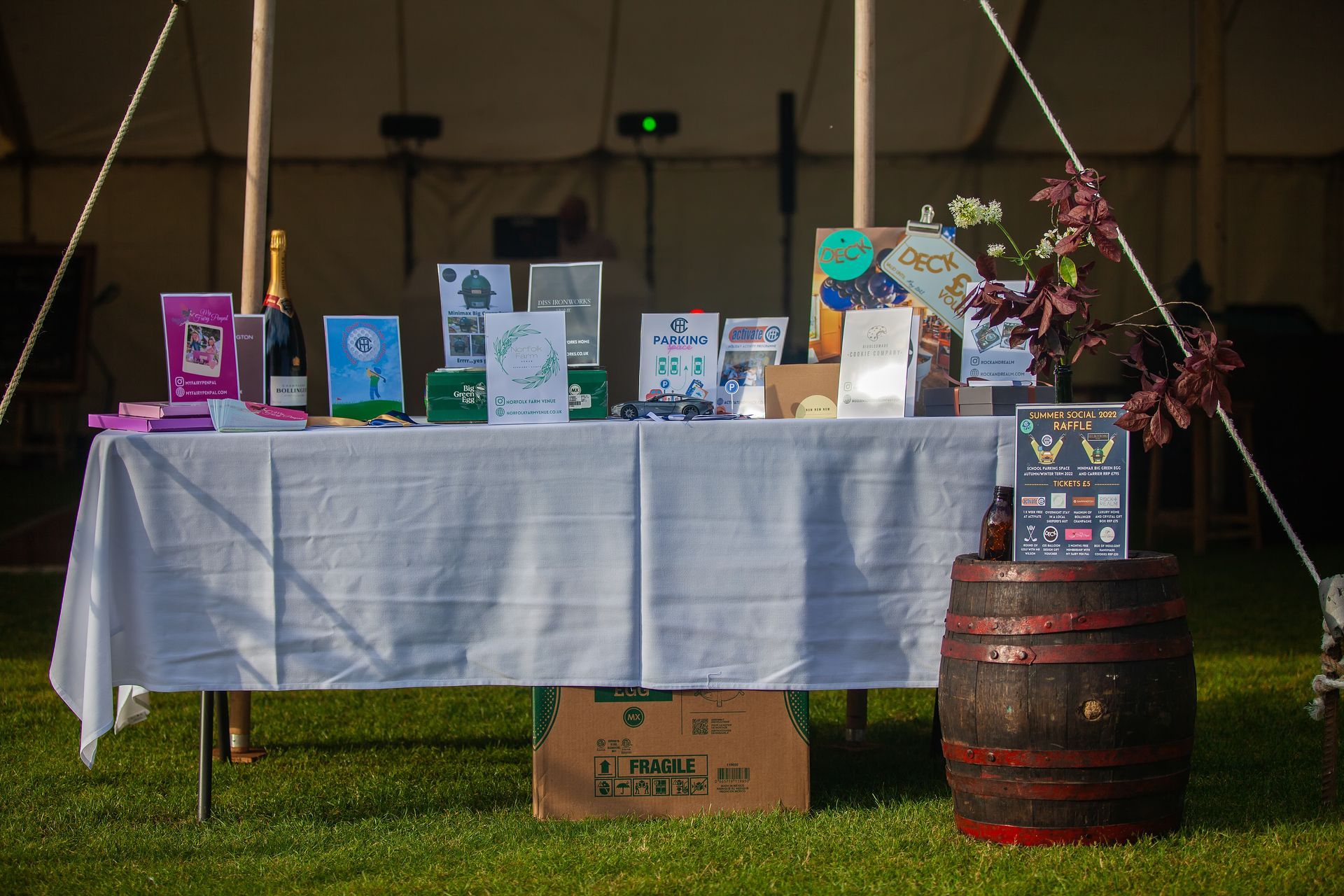 A table with items and a barrel in a tent on grass; items include awards, champagne, and cards.