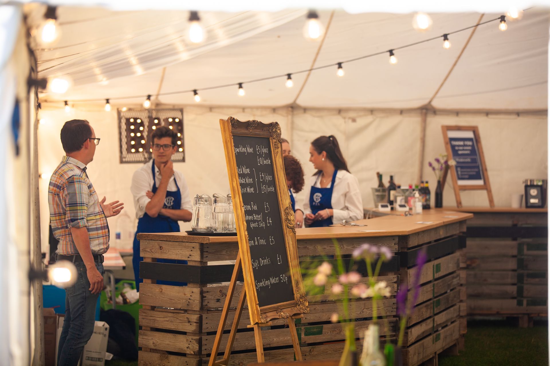 A bar inside a tent. People interact at the counter. String lights hang above.