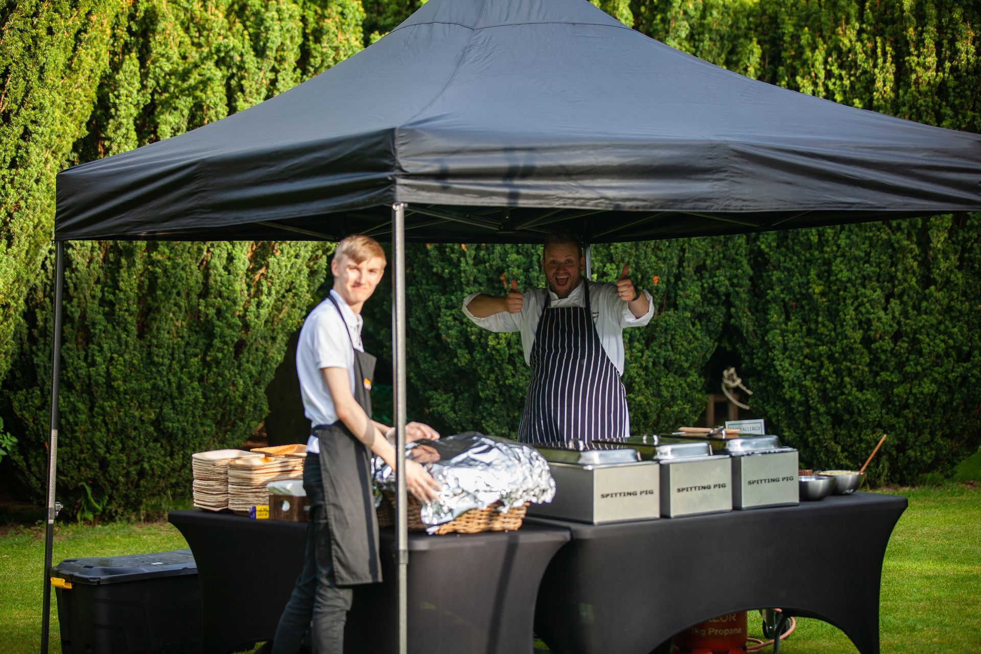 Two caterers at an outdoor event under a black tent, preparing food on a table covered in black cloth.