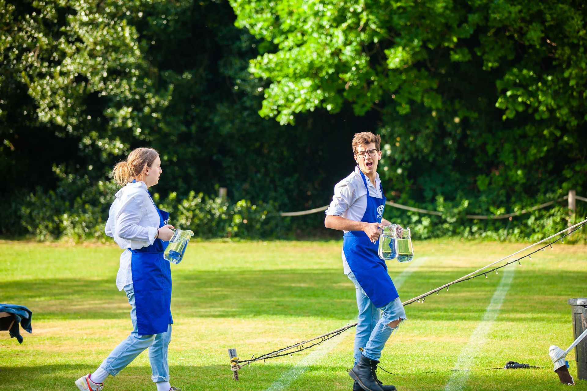 Two people in blue aprons and jeans on a grassy field, likely serving at an outdoor event.