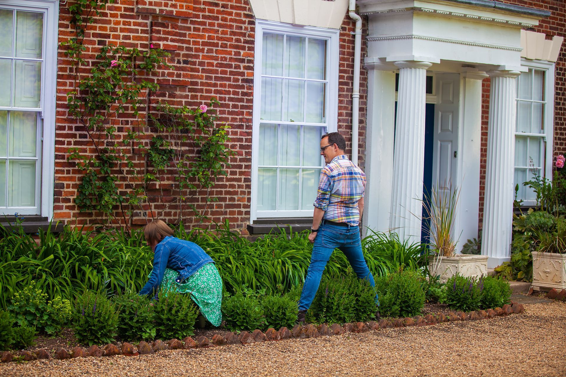 Two people gardening in front of a brick house. One kneels, the other stands.