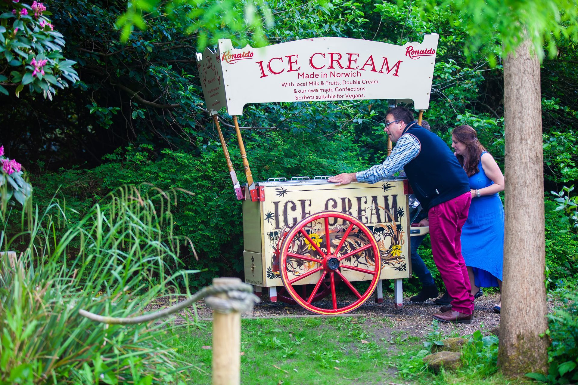 Man and woman by vintage ice cream cart in a garden setting.