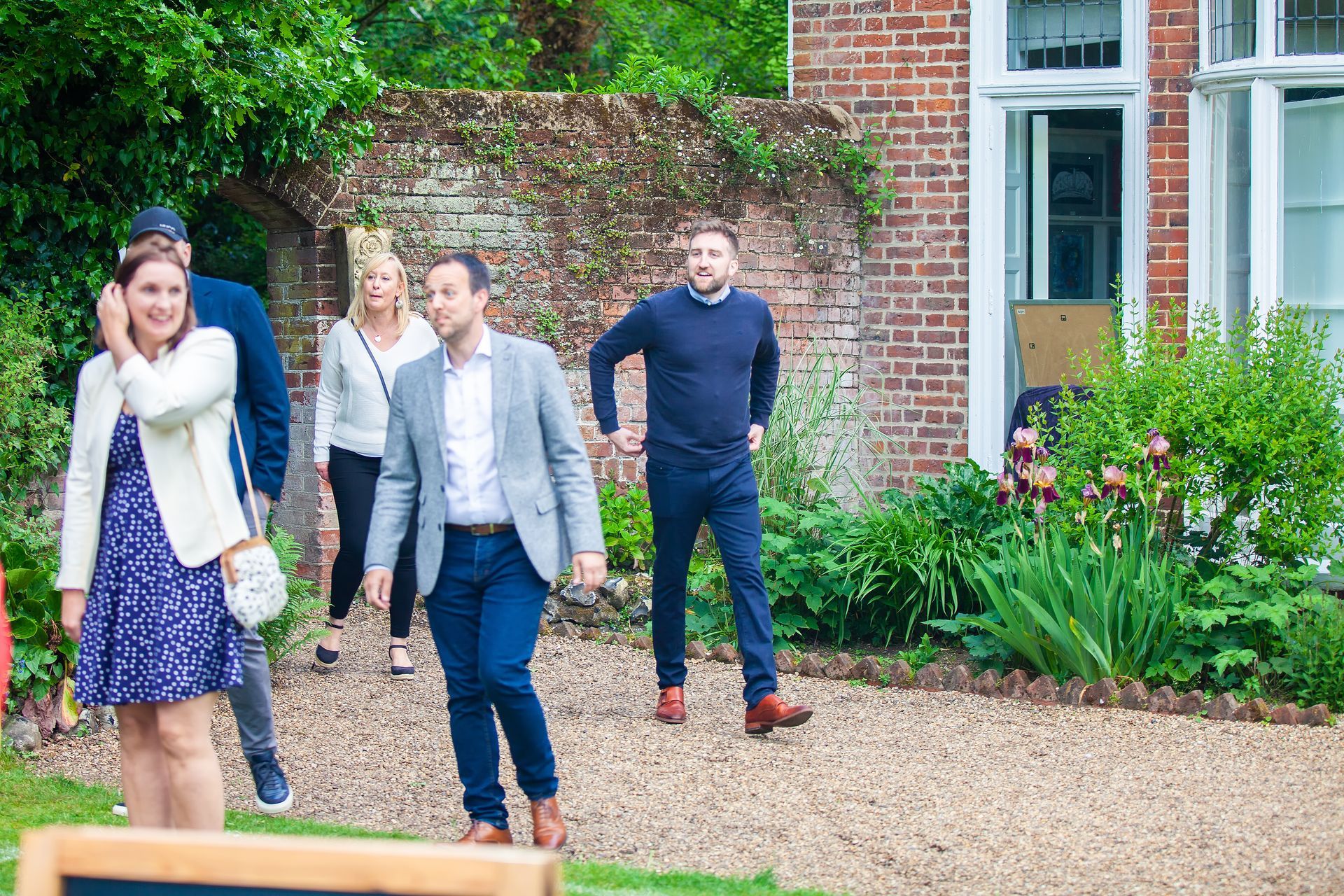Group of people walking outdoors near a brick building and garden; some are smiling.
