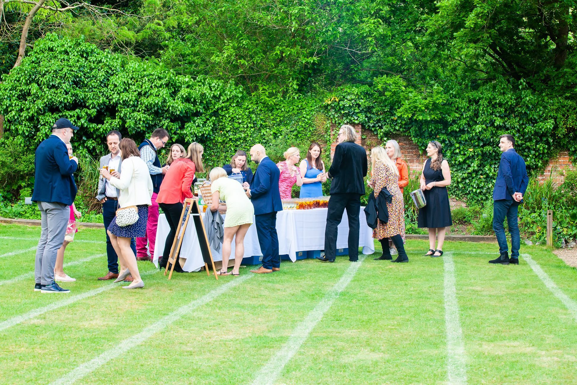 People gathered around a table on a grassy lawn; a party, food and drinks, in a garden setting.