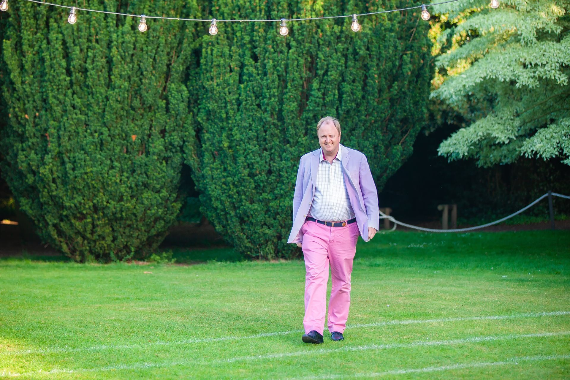 Man in pink pants and lavender jacket walks on grass under string lights.