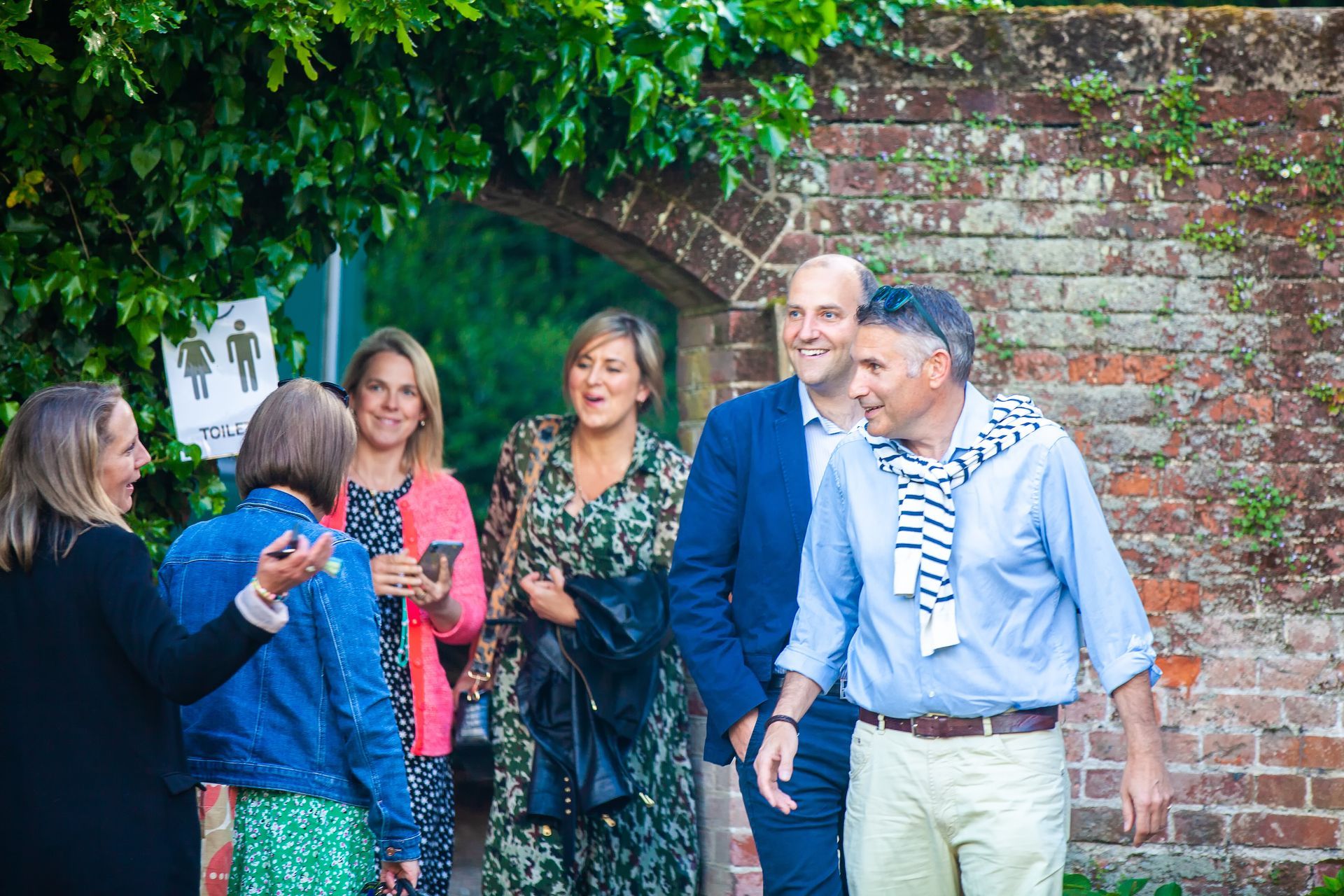 Group of people, including a man in a blue suit, walk through a brick archway, laughing in an outdoor setting.