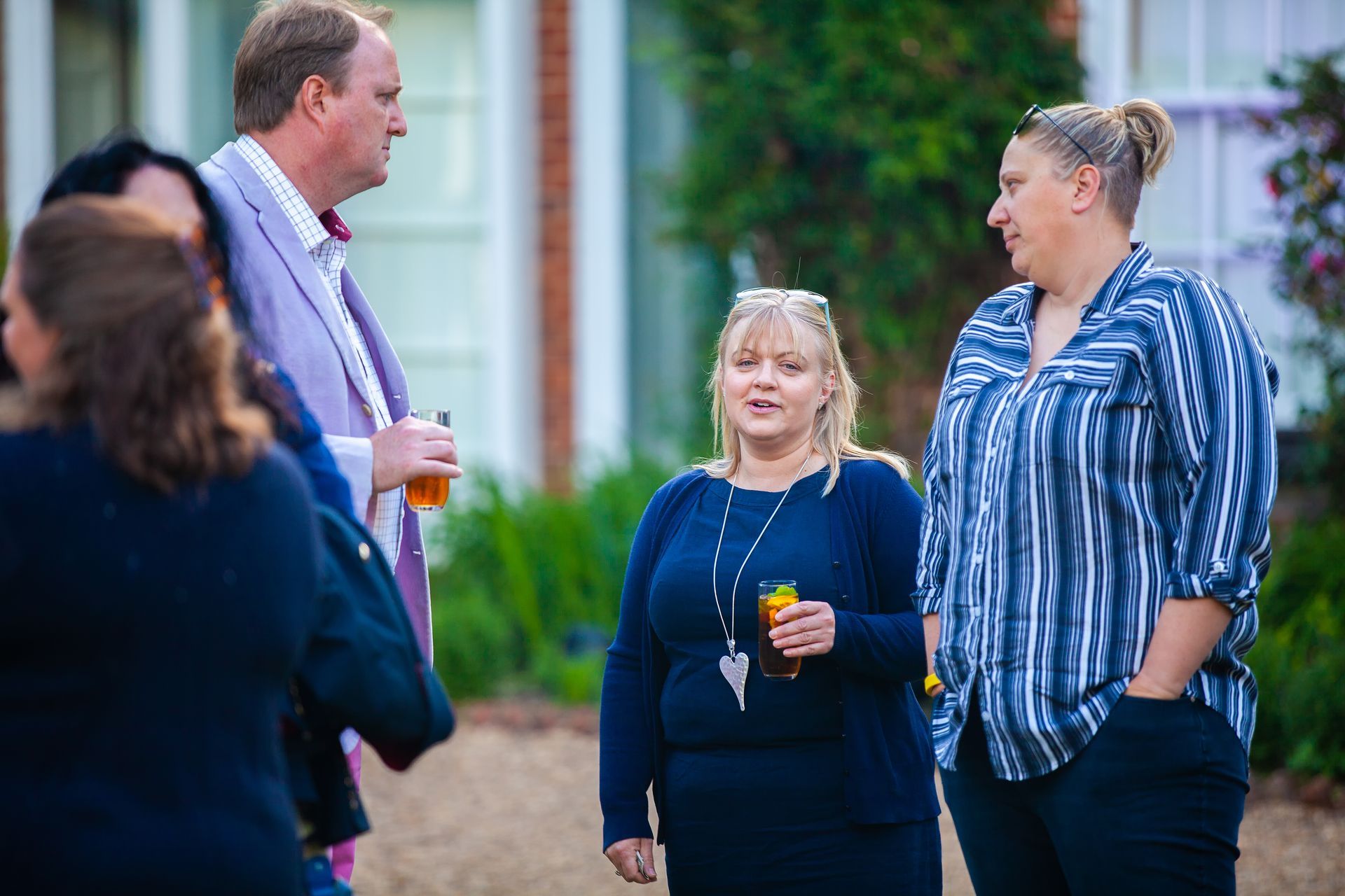 Group of people chatting outdoors, holding drinks. A woman in a blue sweater is in focus.