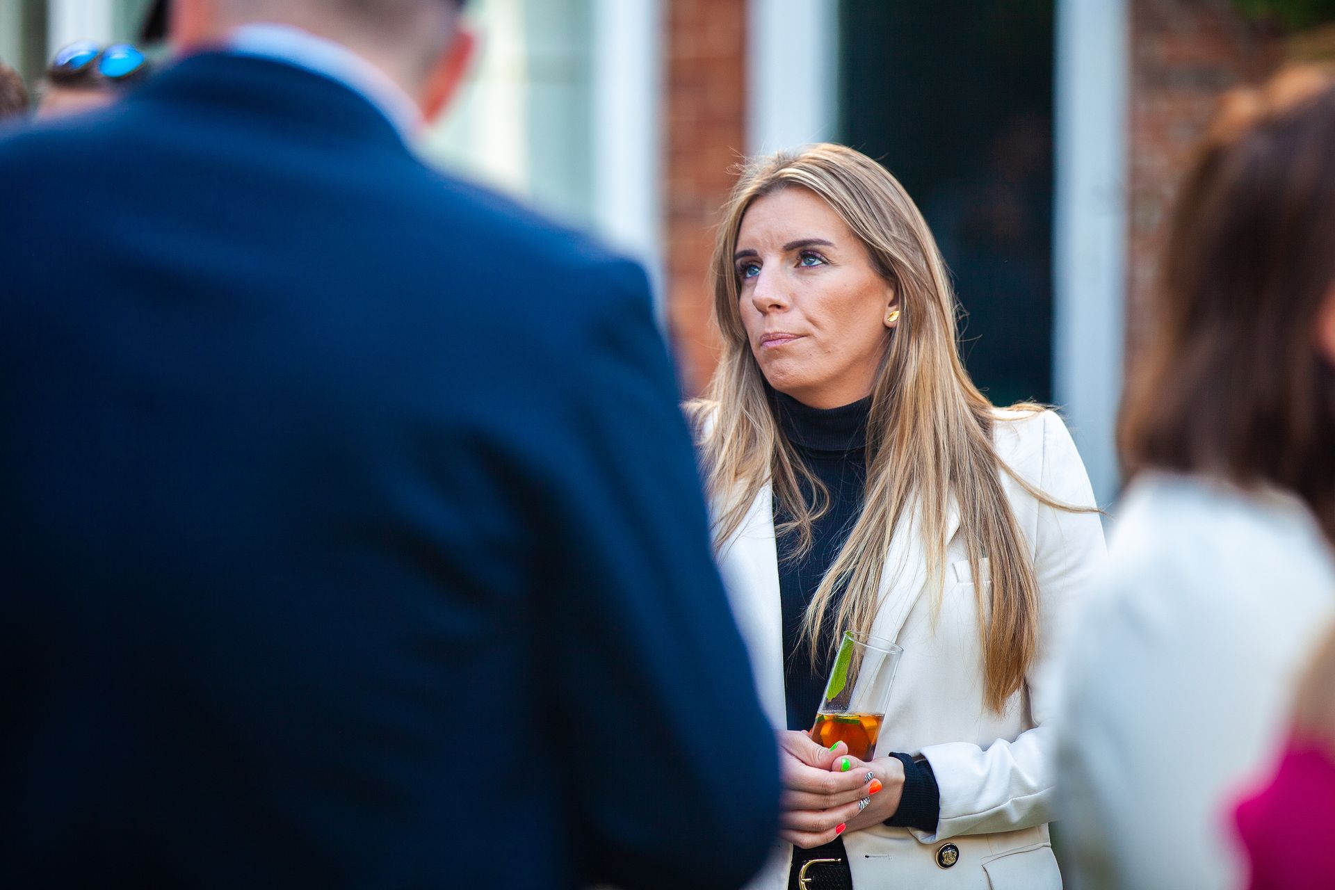 Woman in white blazer holding a drink, looking thoughtful, with a man in a suit and other people nearby.
