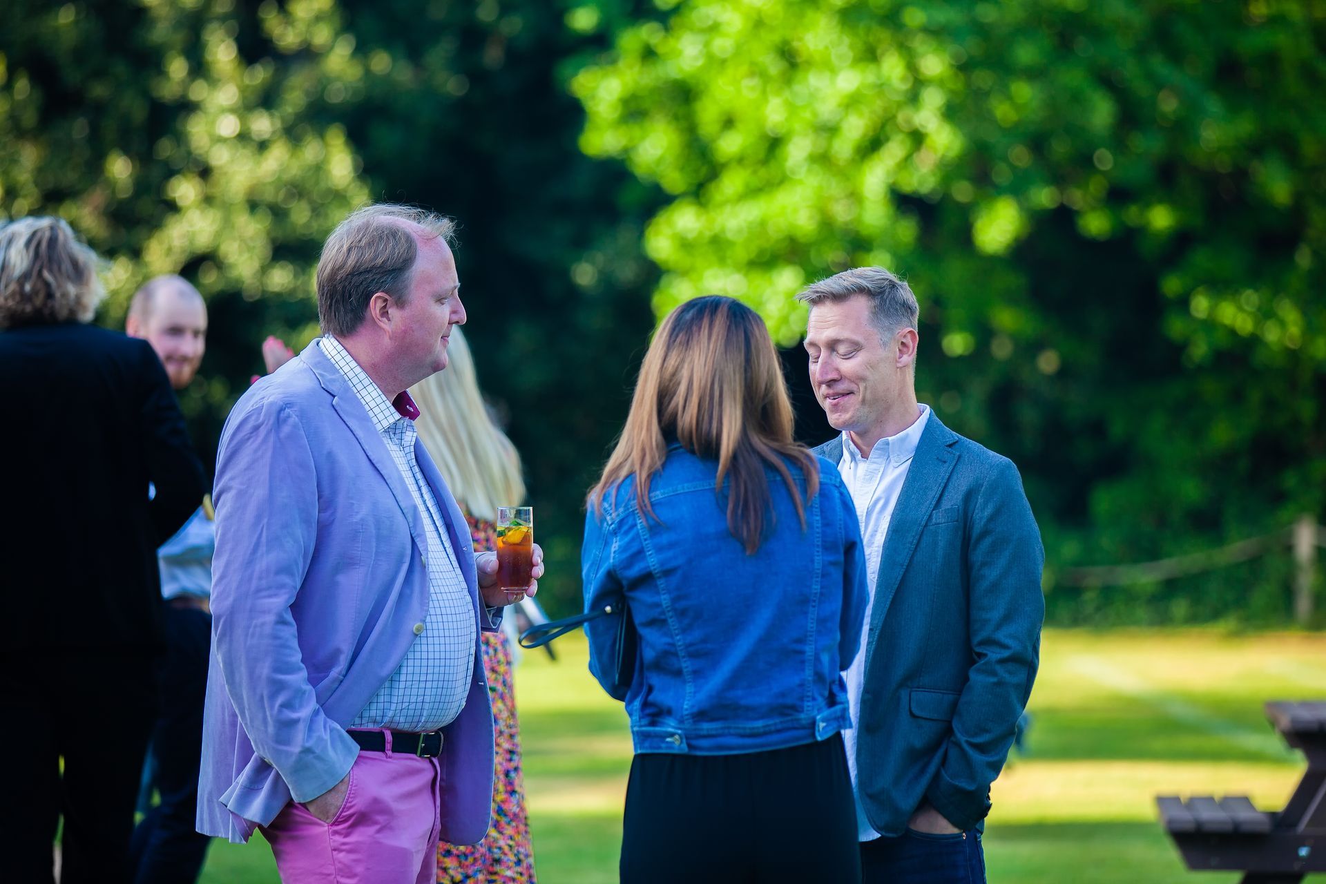 Group of people chatting outdoors, man in pink pants and lilac jacket, sunny day.
