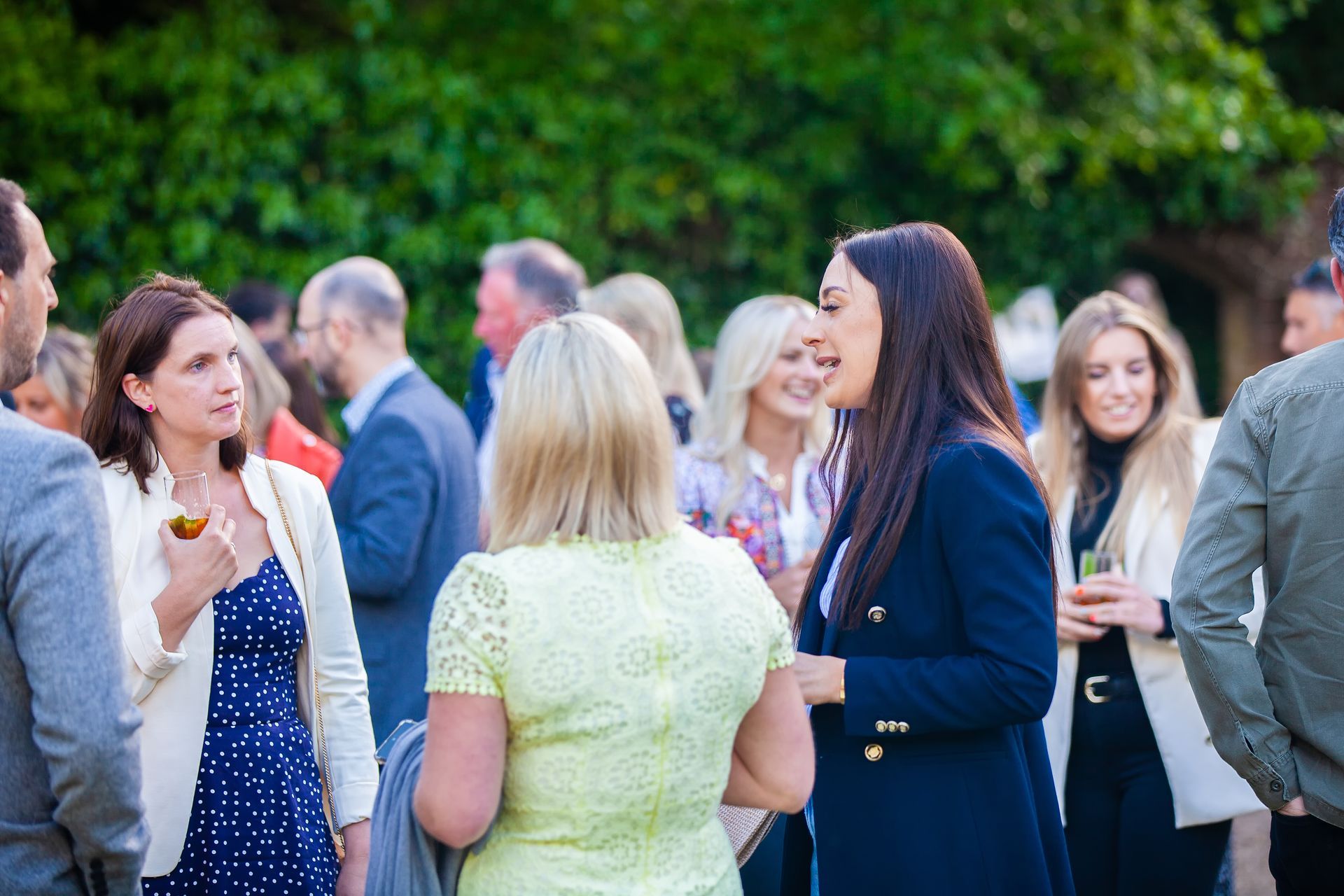 Group of people socializing outdoors, some holding drinks; women in focus, smiling.