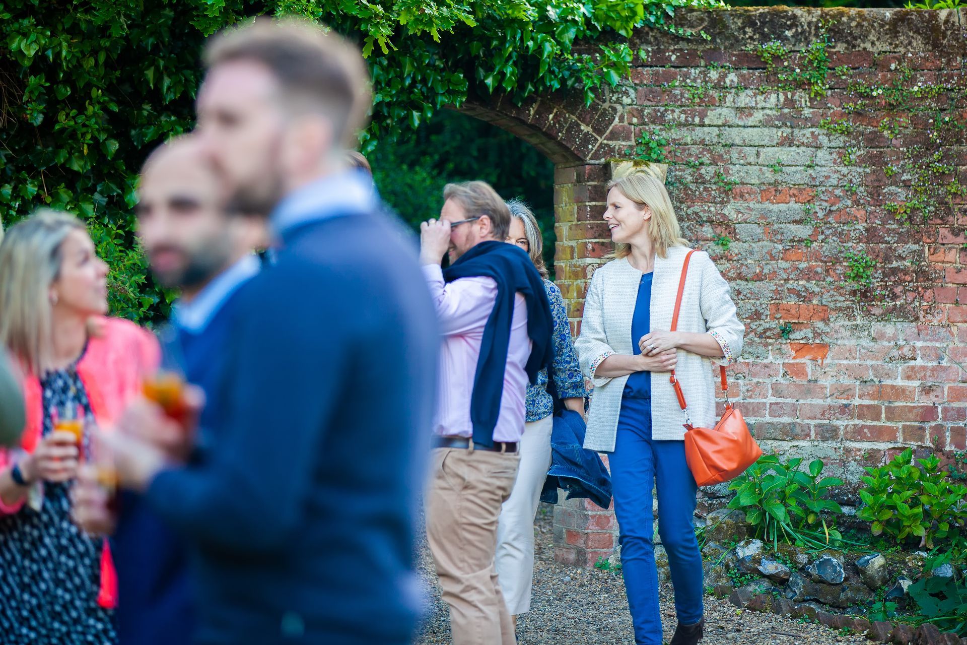 People at an outdoor event, woman in white jacket and orange bag walks past a brick wall, other people socializing.