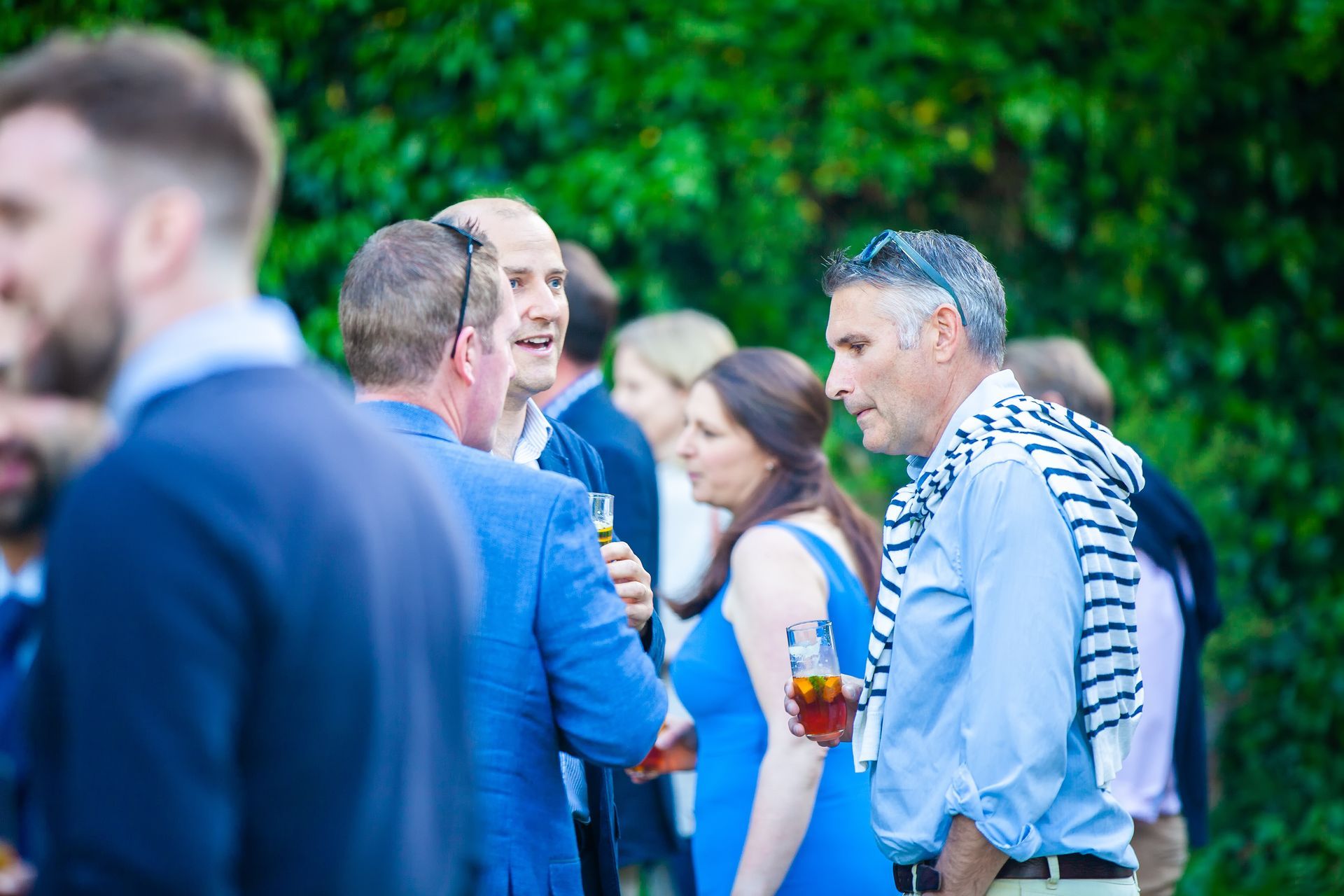 Group of people socializing outdoors, some holding drinks.  Green foliage in the background.