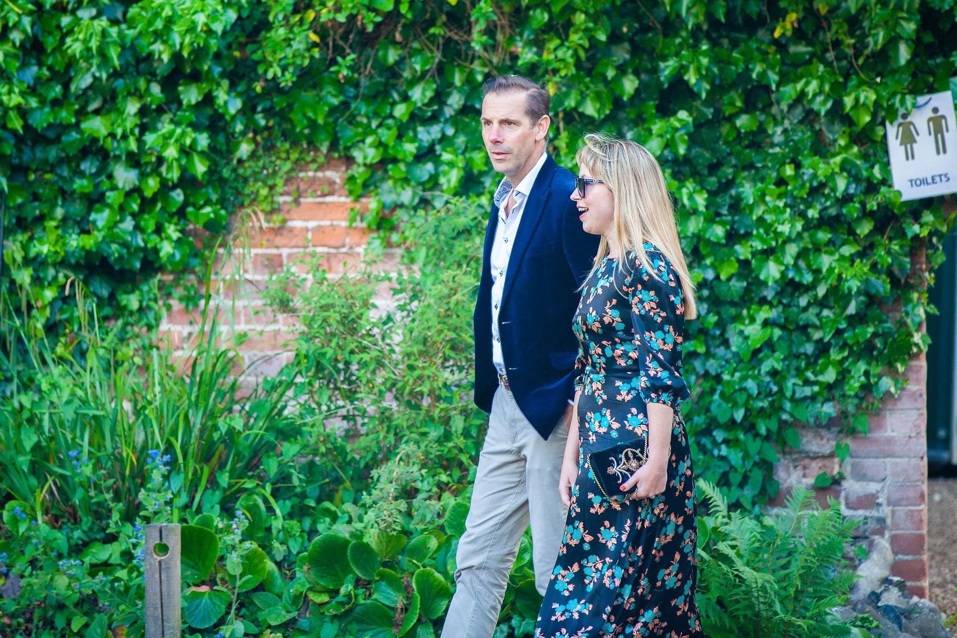 Man and woman walking past greenery; woman in floral dress, man in blazer.