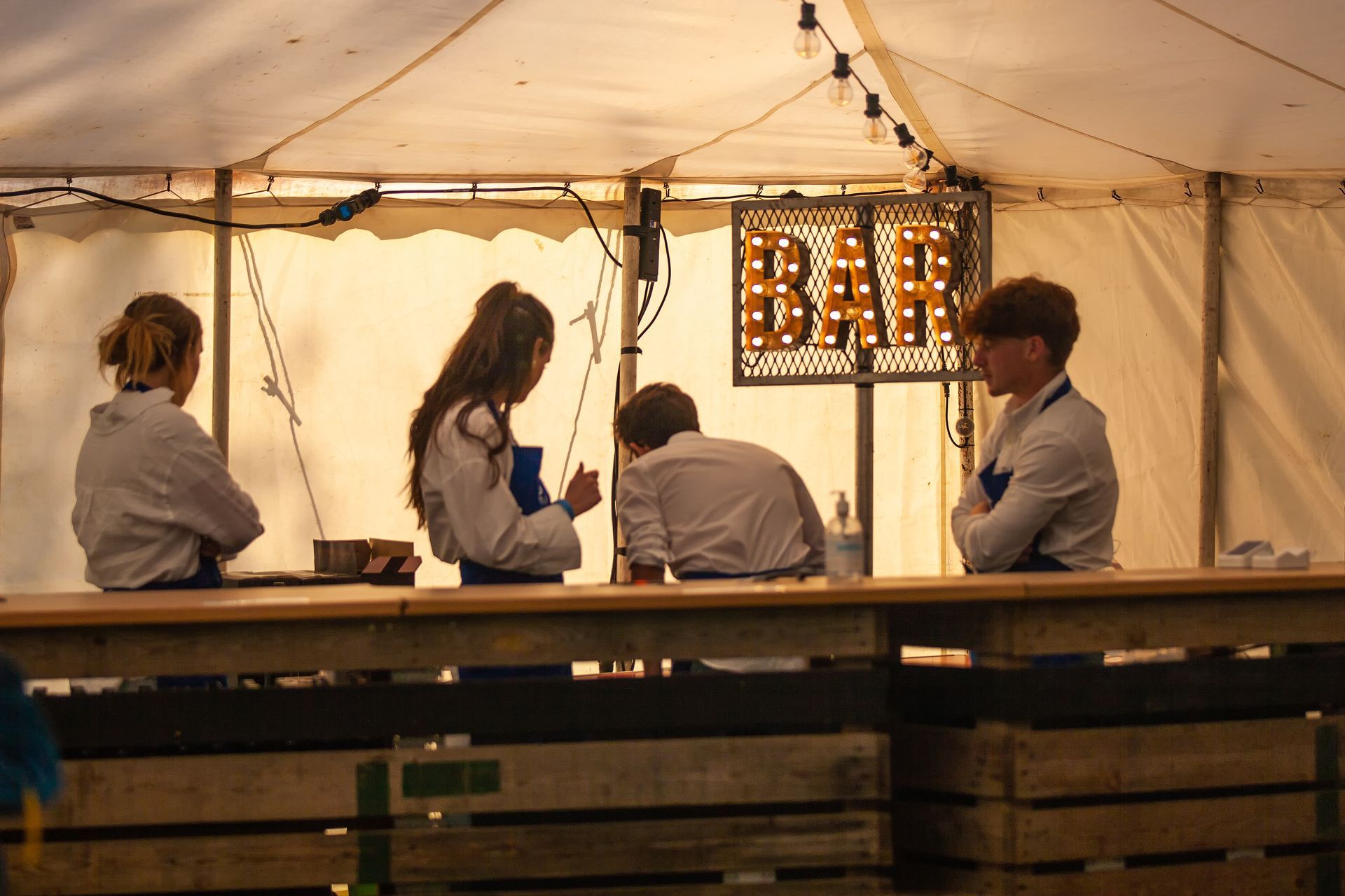 Four young people behind a bar in a tent, wearing aprons. 