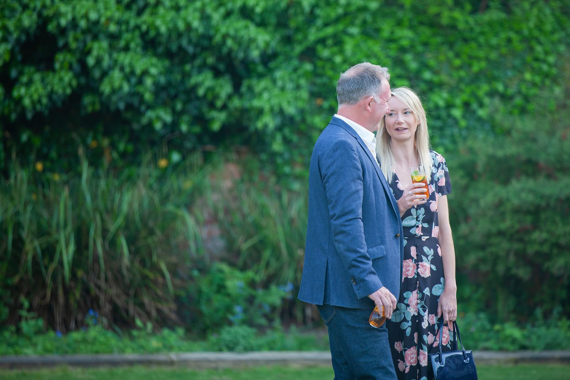 Man and woman conversing outdoors; woman holds drink; both are near greenery.