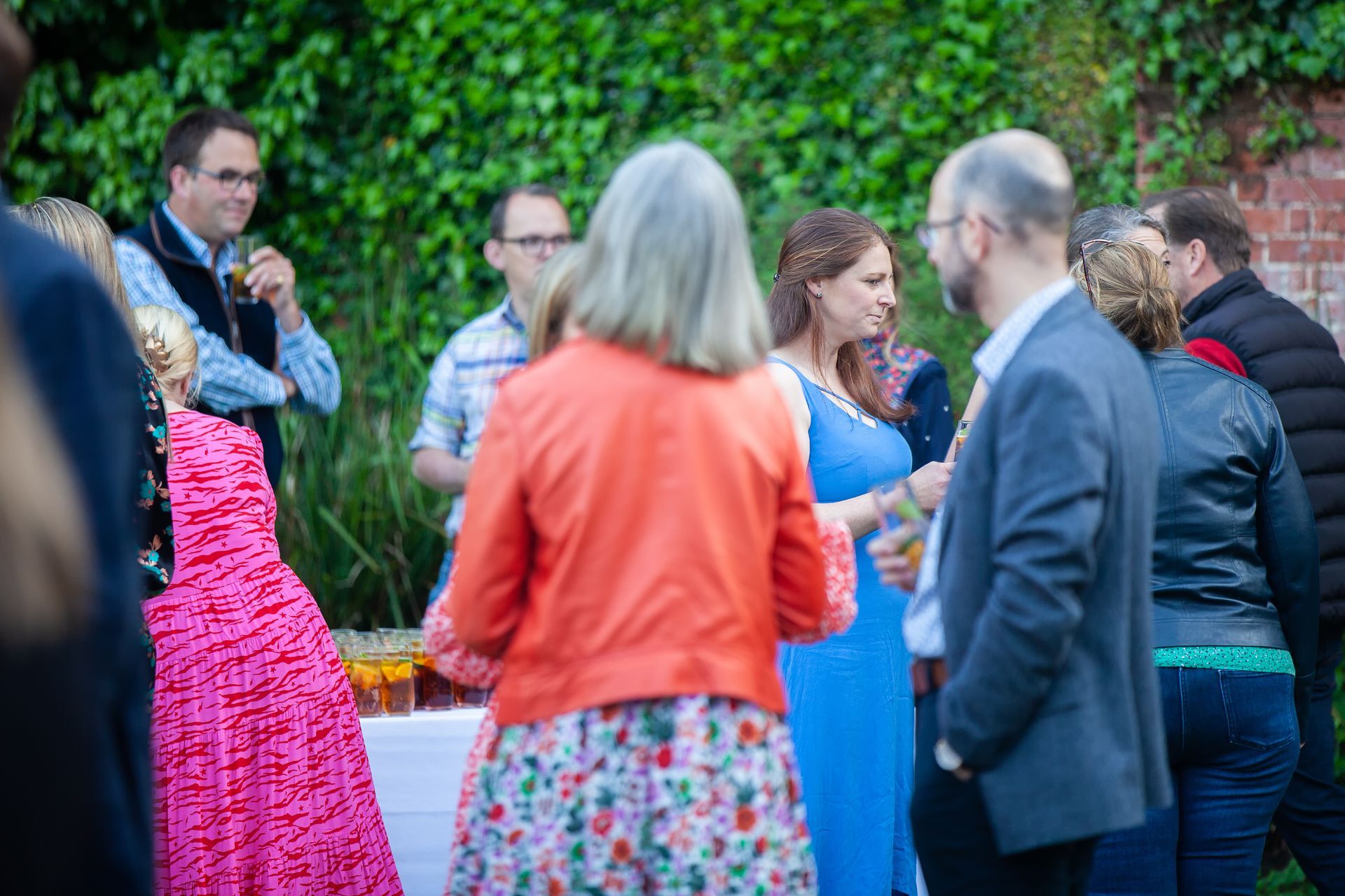 People at outdoor gathering, socializing. Woman in orange jacket, pink dress, and blue dress visible.