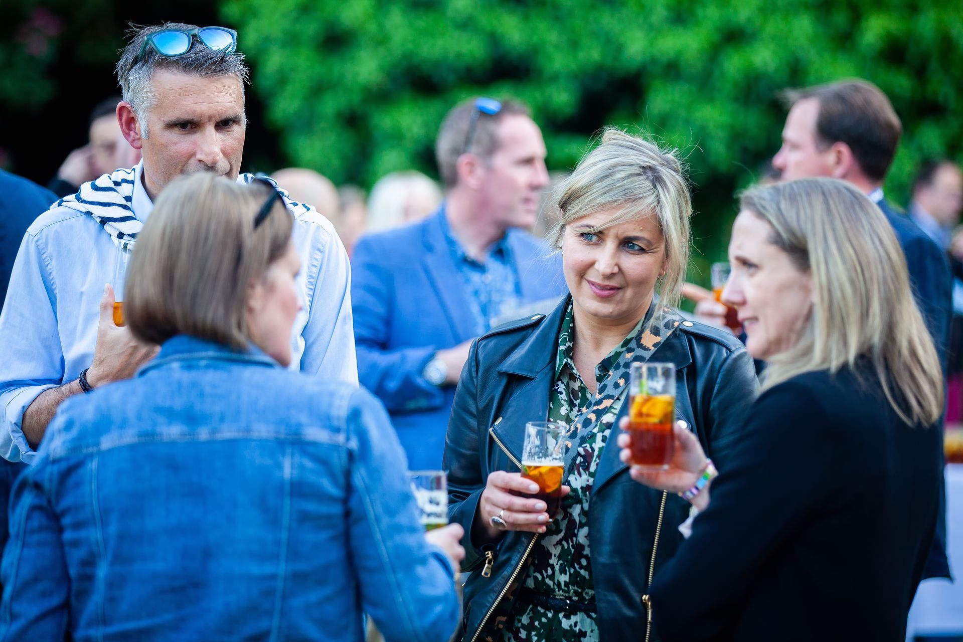 Group of people chatting and drinking beverages at an outdoor gathering; trees in the background.