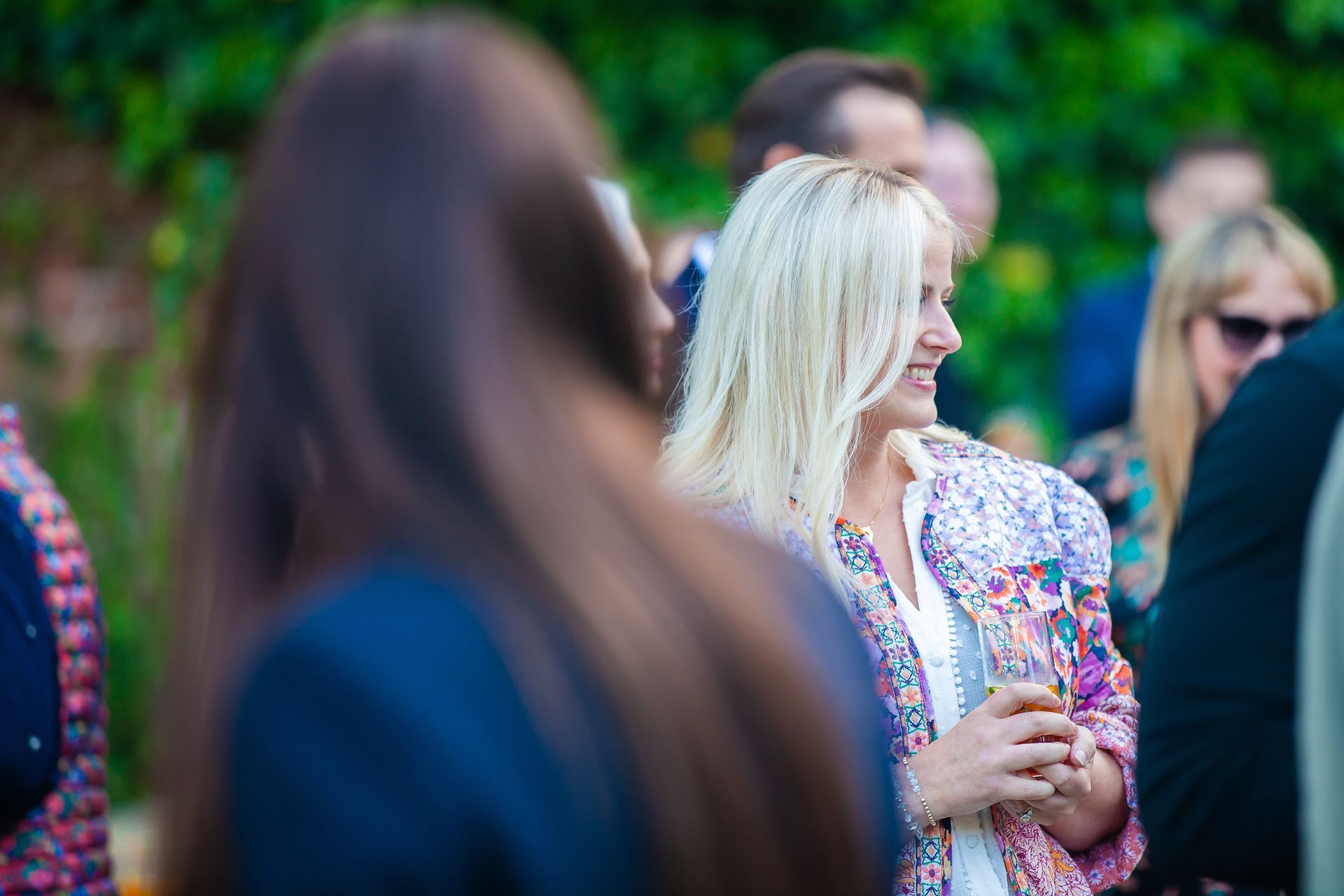 Blonde woman in floral jacket smiles, surrounded by people outdoors.