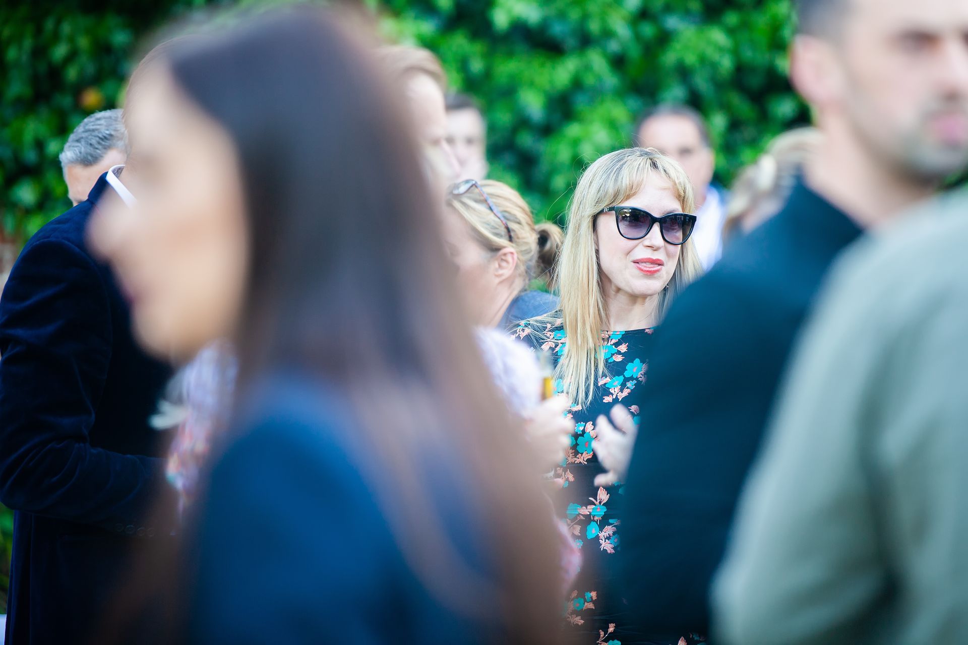 Woman in sunglasses smiles at an outdoor gathering, surrounded by other people and greenery.