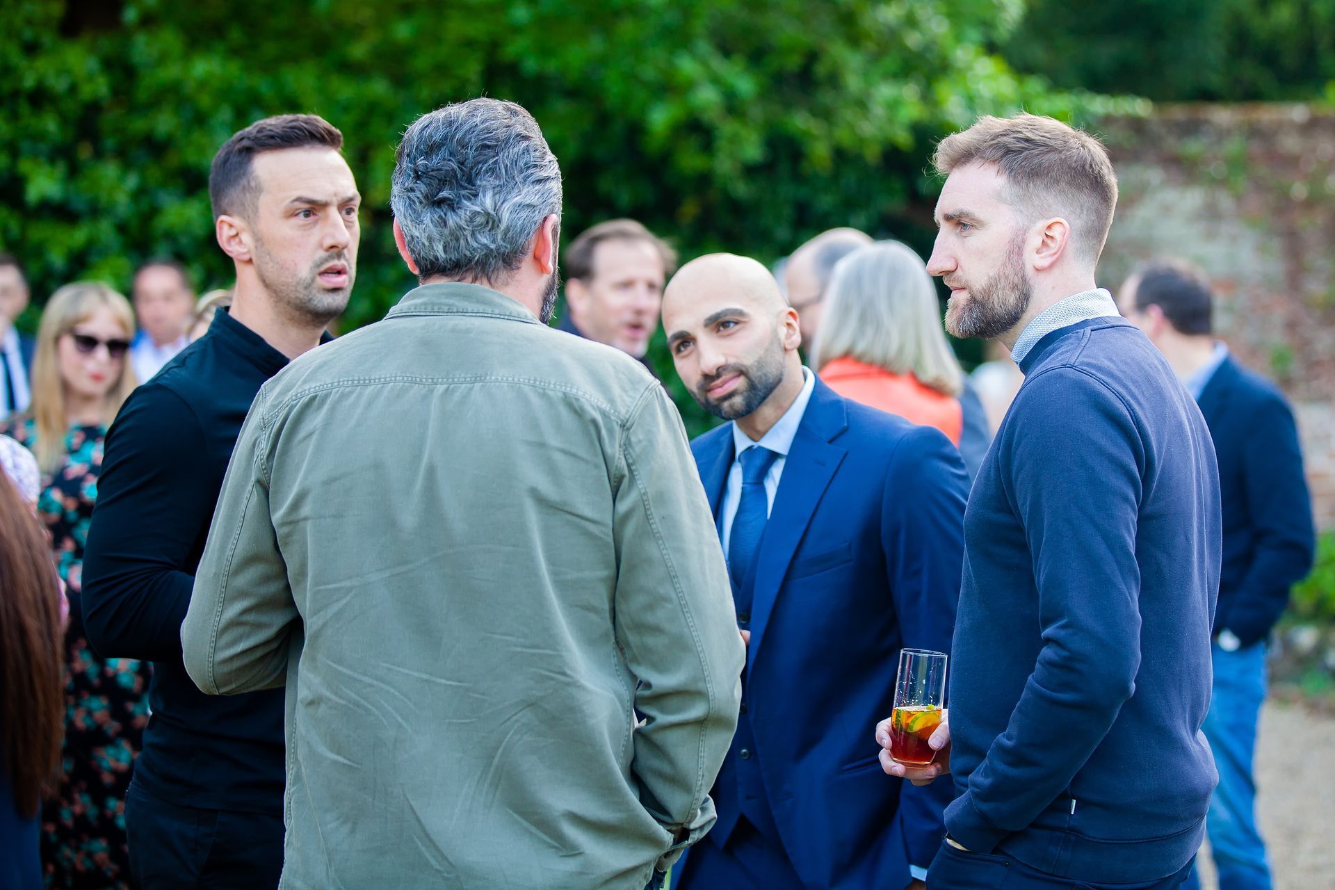 Men in an outdoor gathering, conversing and holding drinks, dressed in casual and formal attire.